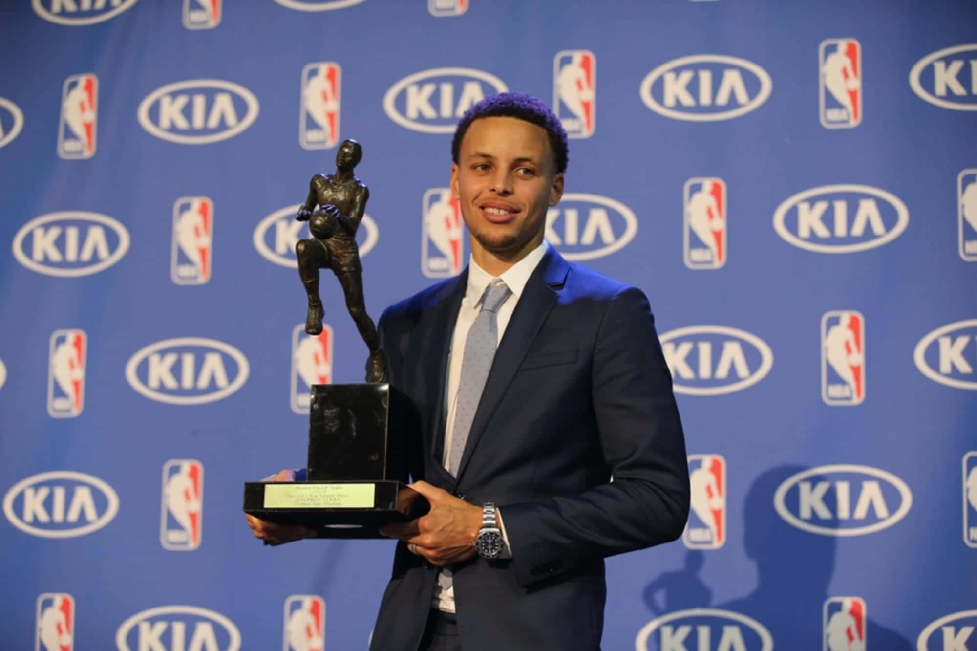 steph curry 桌布 壁紙 — 史提芬居里微笑著，在帶有NBA和Kia標誌的藍色背景前手持籃球獎盃。