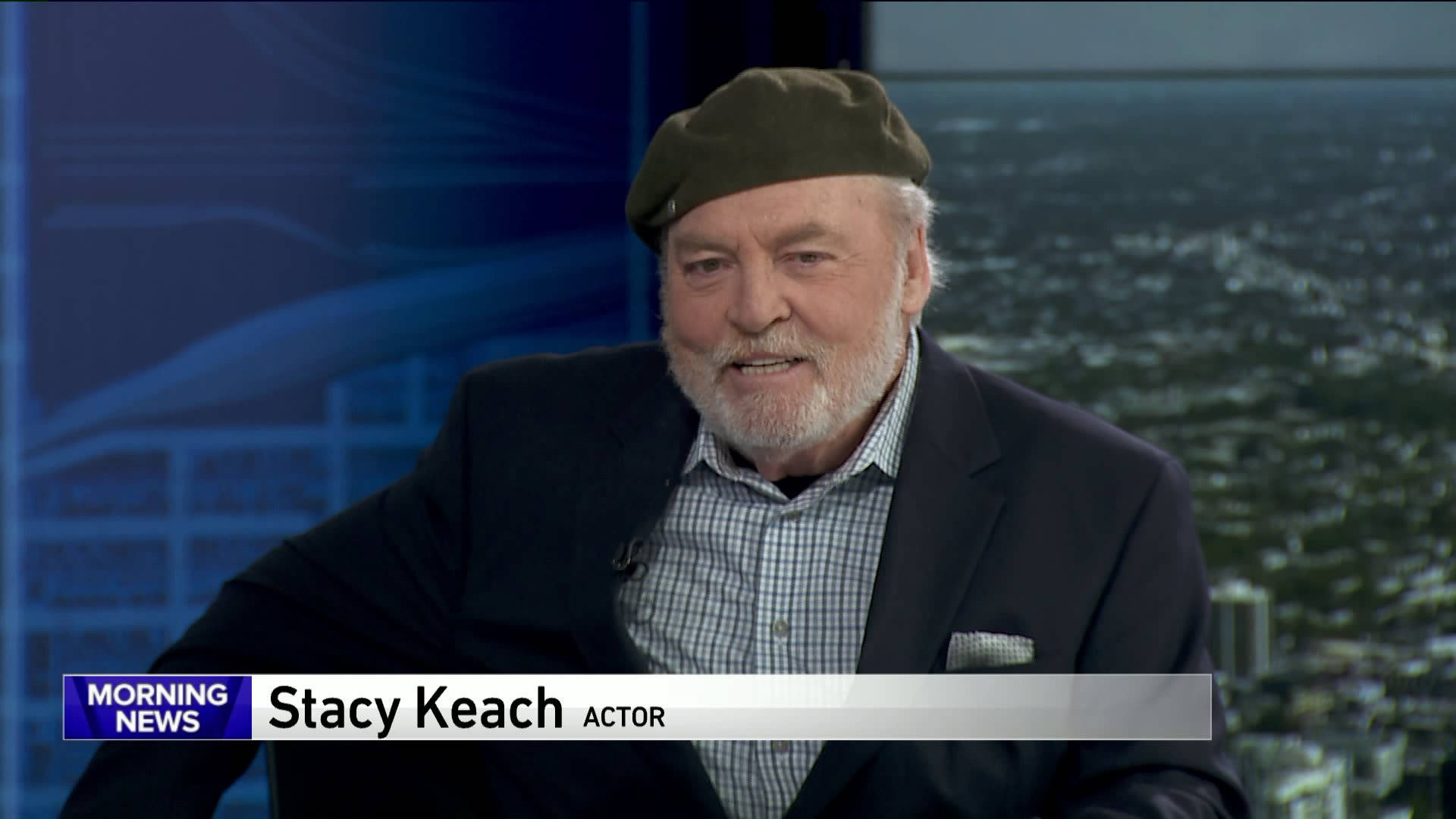演員 Stacy Keach 在工作室接受訪問，身後是城市景色。