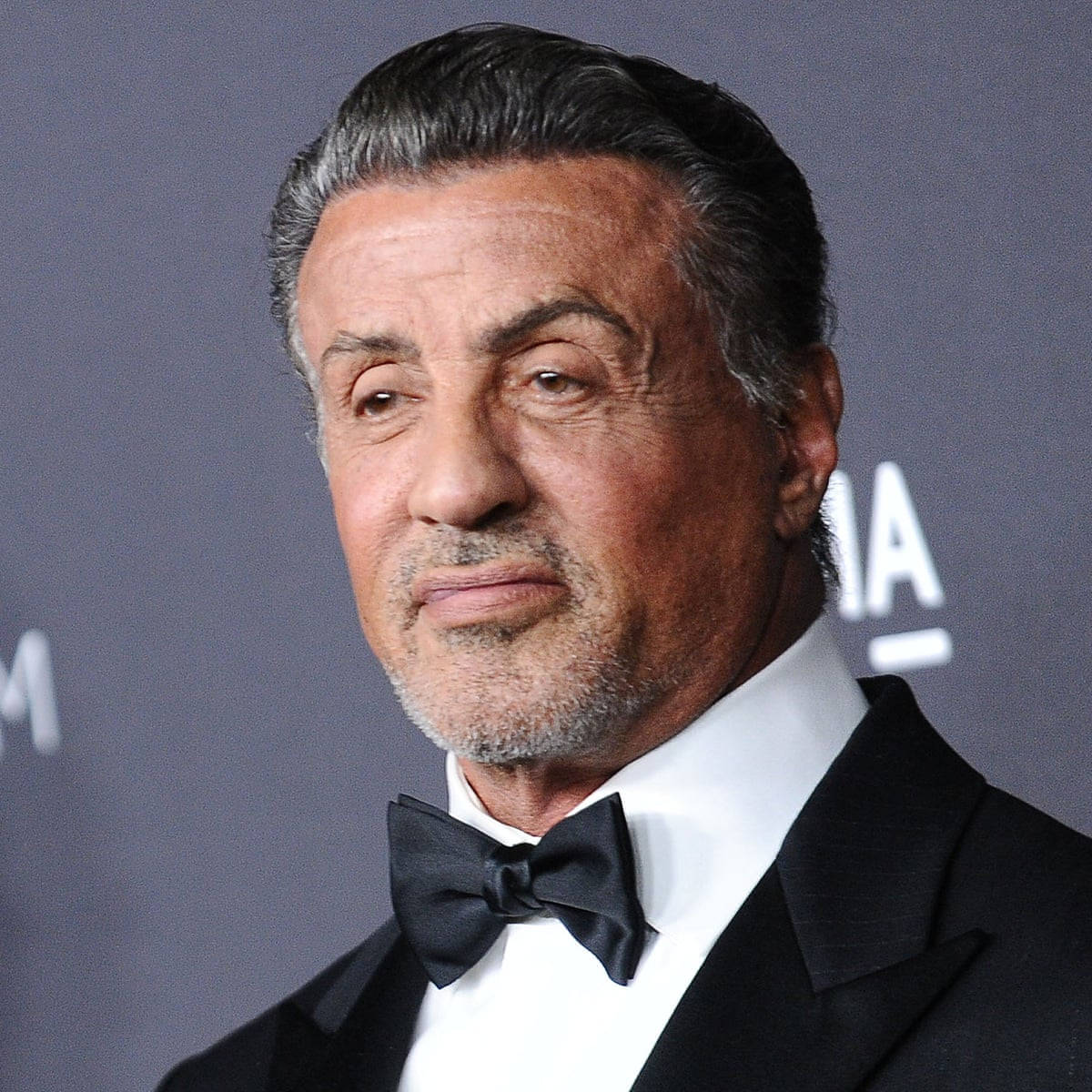 史泰龍穿燕尾服配黑領結 壁紙 — Sylvester Stallone 身穿黑色修身燕尾服和領結，在灰色活動背景前擺出嚴肅的表情。