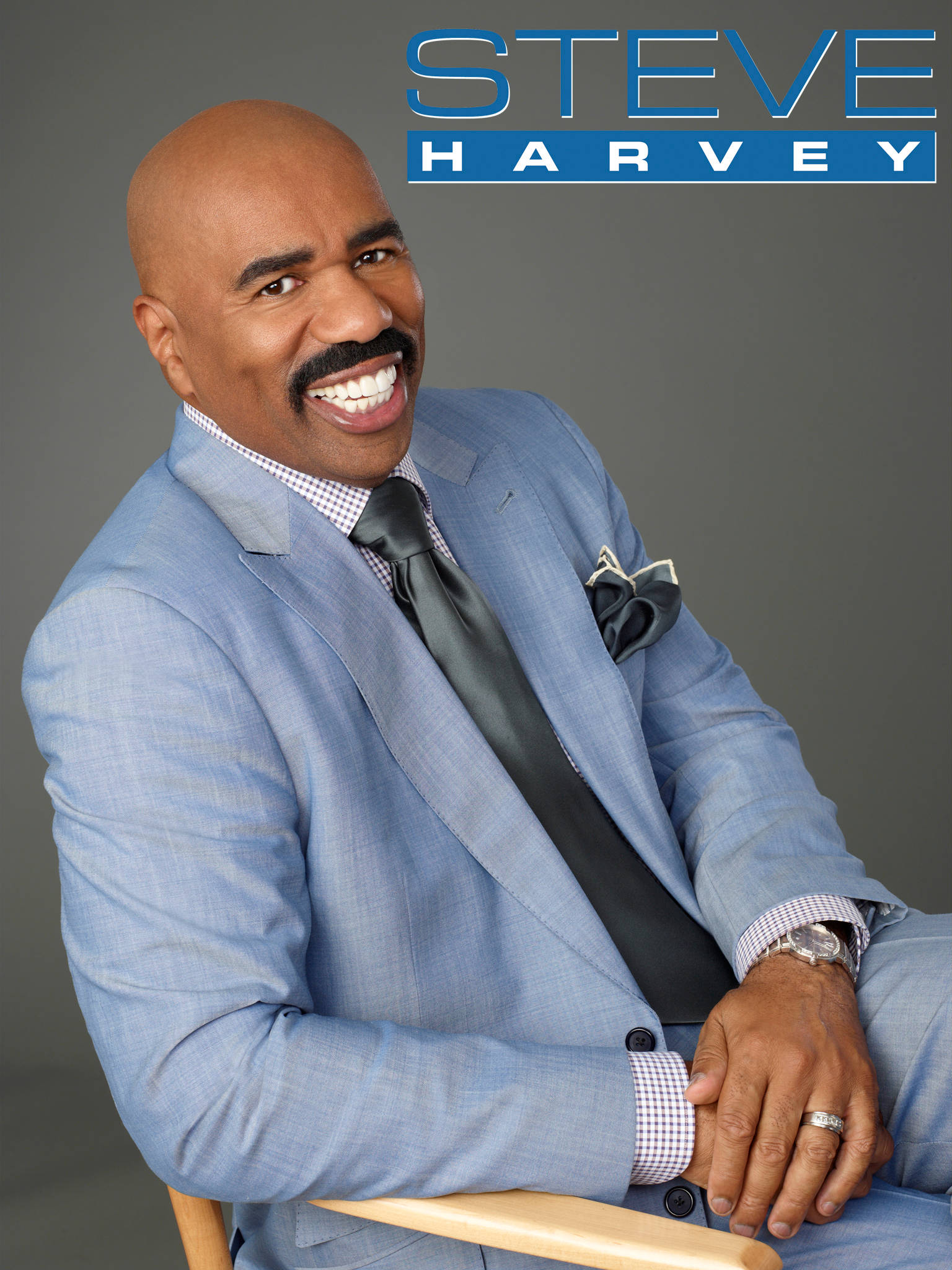 史蒂夫·哈維坐著 壁紙 — Steve Harvey 身穿亮麗淺藍色西裝，在灰色中性攝影棚背景下展露燦爛笑容。