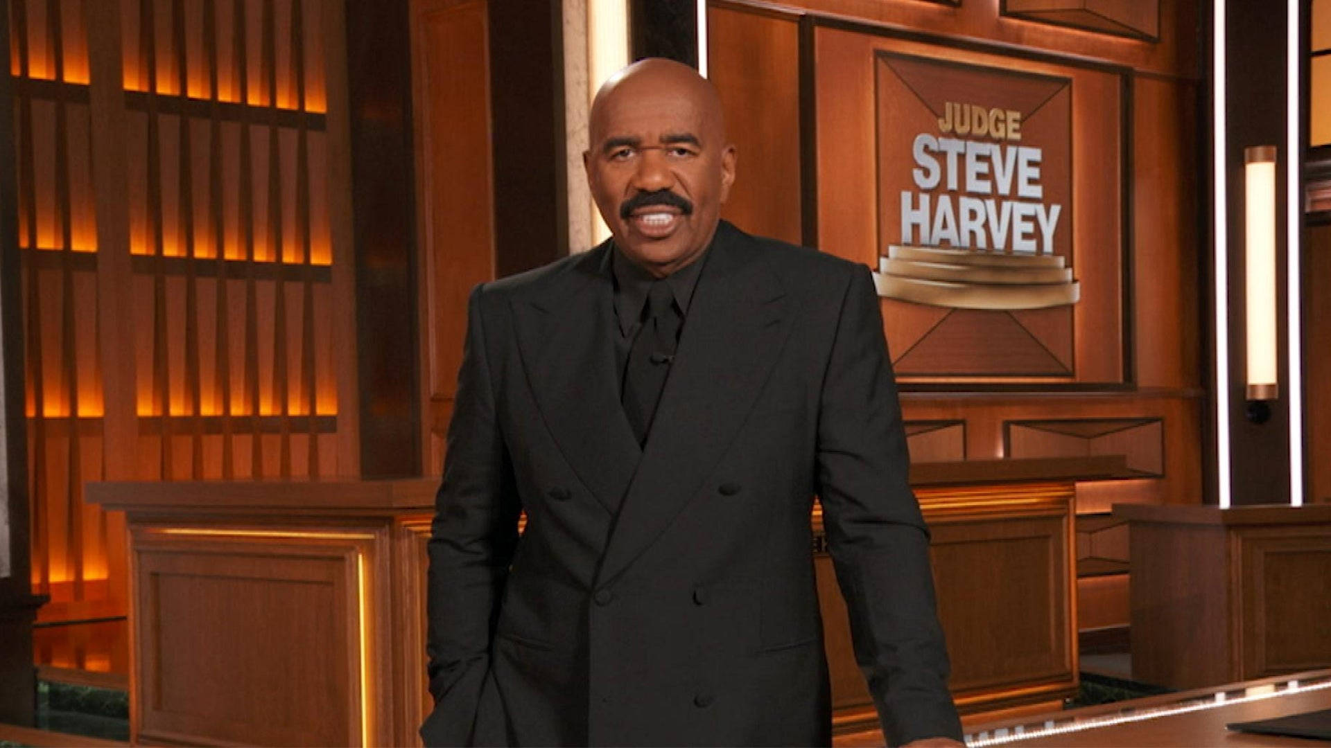 史蒂夫·哈維搭配棕色背景 壁紙 — Steve Harvey 身穿黑色西裝，自信地站在燈光溫暖的木製舞台上，身後有「Judge Steve Harvey」標誌。