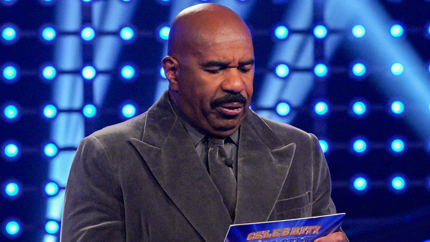 Steve Harvey 穿著灰色西裝，在明亮的舞台上拿著一張藍色卡片。