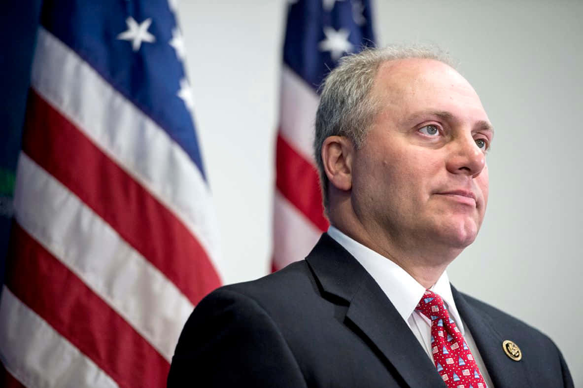 史蒂夫·斯卡利斯身後有美國國旗 壁紙 — 一名身穿深色西裝、繫著紅色圖案領帶的男子，表情嚴肅地站在兩面美國國旗前。