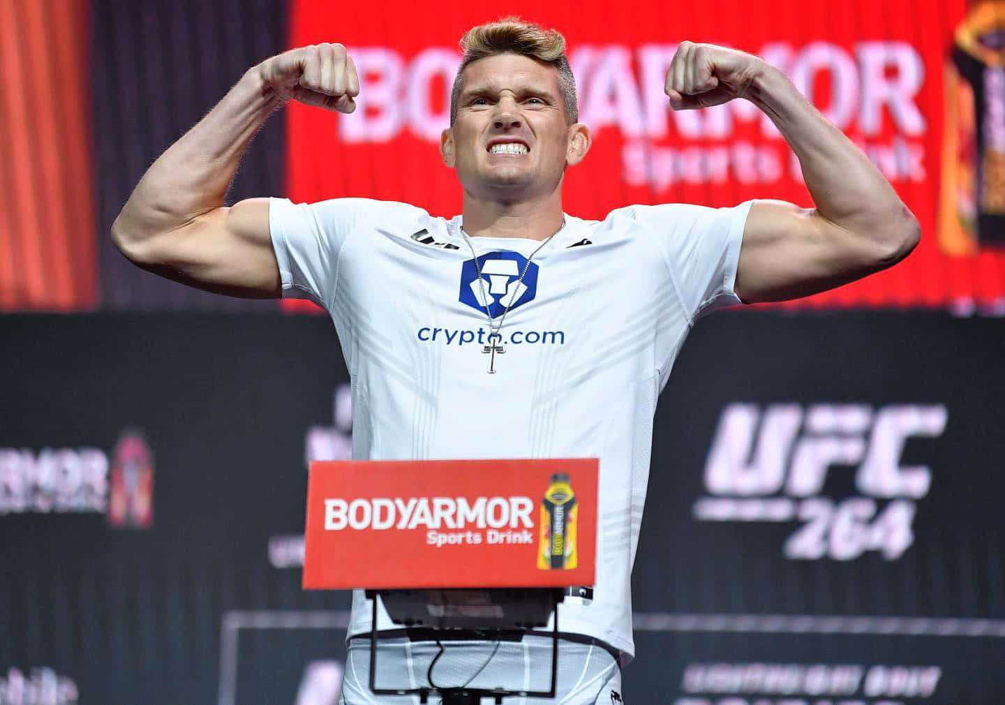 UFC 拳手在稱重活動中彎曲二頭肌，身穿帶有加密貨幣標誌的白襯衫。