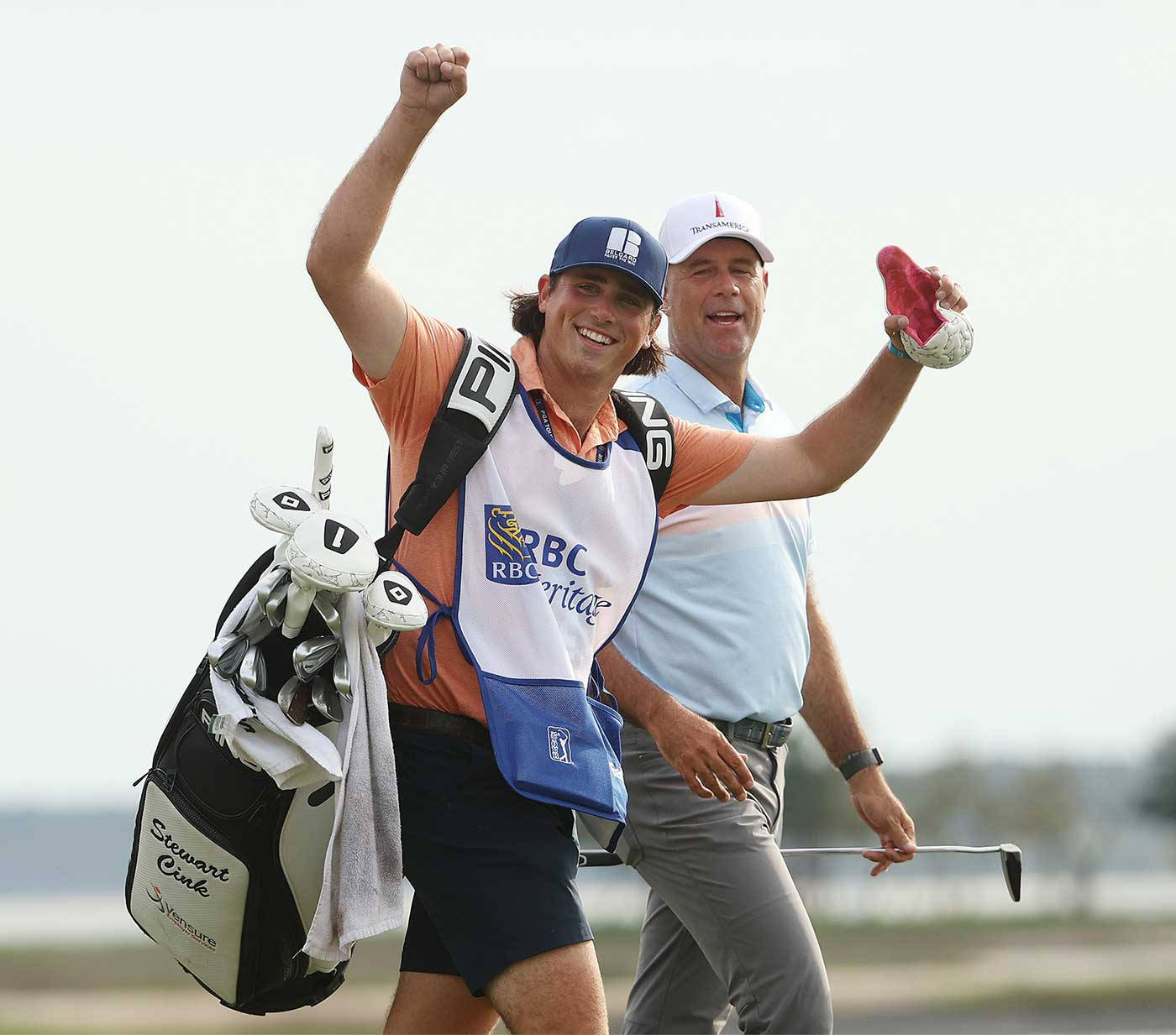 史都華·辛克與兒子漫步球場 壁紙 — 一位身穿RBC Heritage背心的高爾夫球手欣喜若狂地舉起拳頭慶祝勝利，他的球童在旁邊微笑著，手持高爾夫球桿和手套。