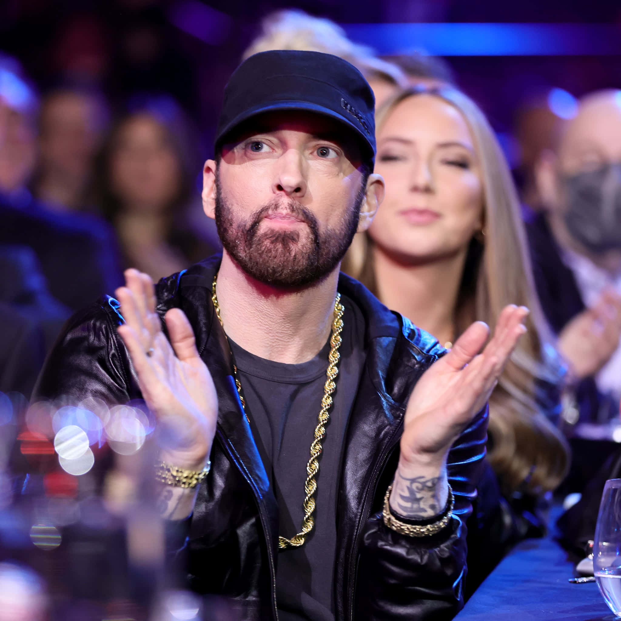 Eminem 桌布 壁紙 — 司儀Eminem在燈光昏暗的活動中熱情鼓掌，戴著黑色帽子、皮褸和金鏈。