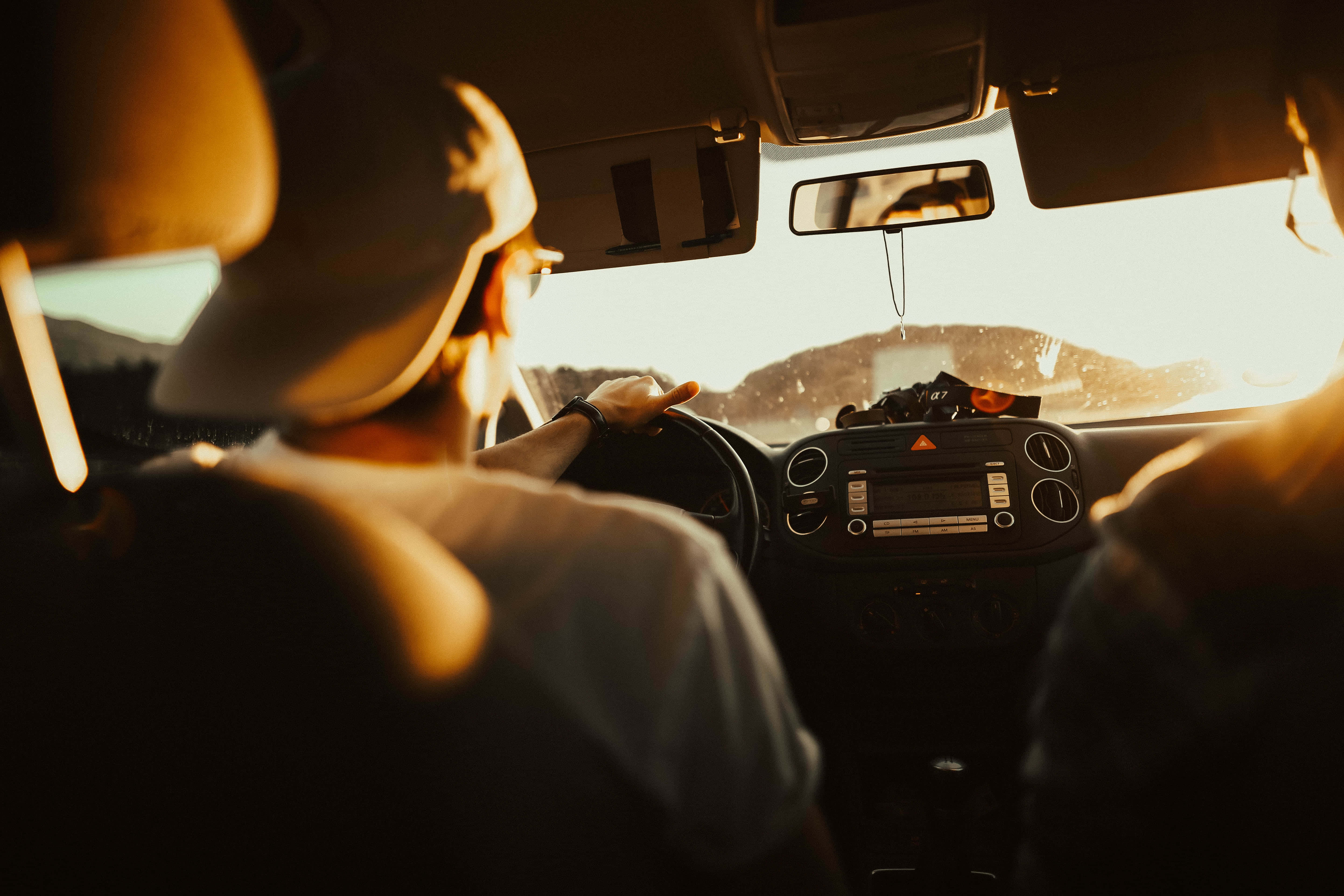 road trip 桌布 壁紙 — 司機戴著帽子在日落時駕駛汽車，溫暖的金色光線充滿車內，透過擋風玻璃可以看到山丘。