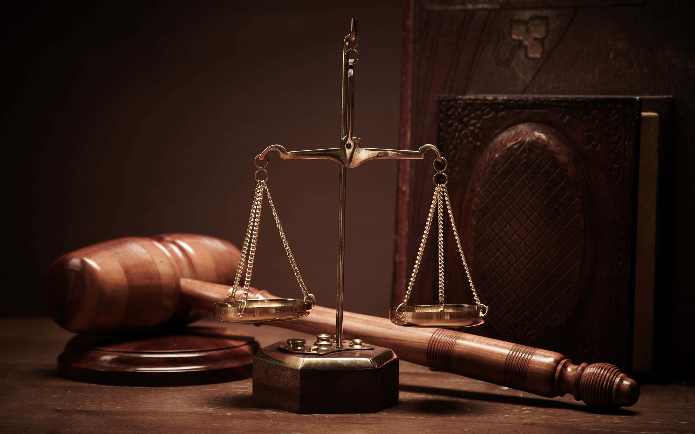 司法法庭正在開庭 壁紙 — 金色正義天秤平衡放在木槌和法律書上，處於溫暖昏暗的燈光中。