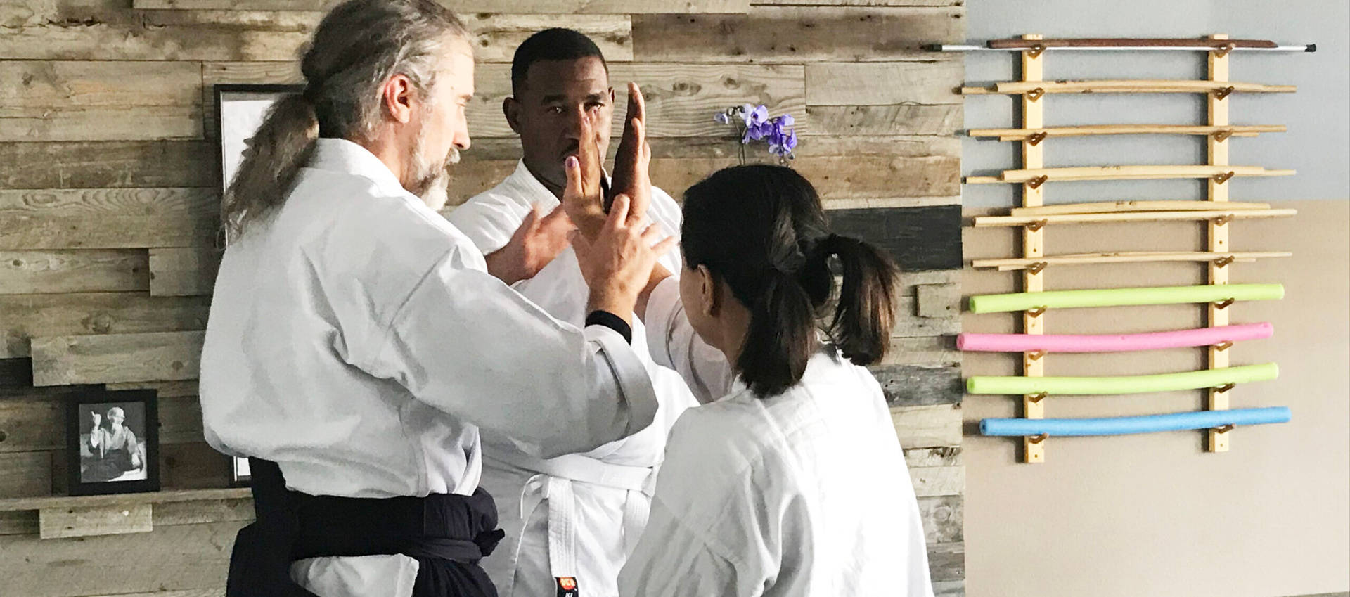 合氣道教練示範二合技 壁紙 — 三位身穿白色道服的武術家，在質樸的木牆前，用色彩繽紛的訓練棍練習精準的手部技巧。
