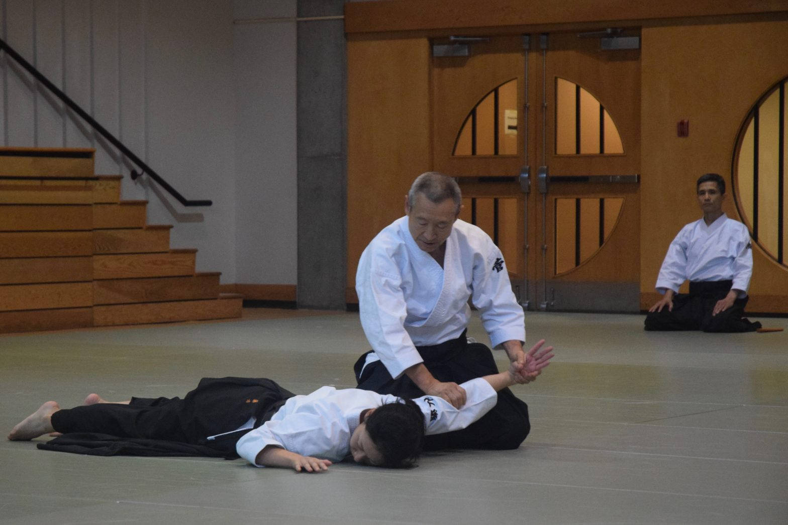合氣道練習者展示二見技 壁紙 — 一位年長的武術教練在一個有木門和樓梯的道場中，向學生演示纏鬥技巧。