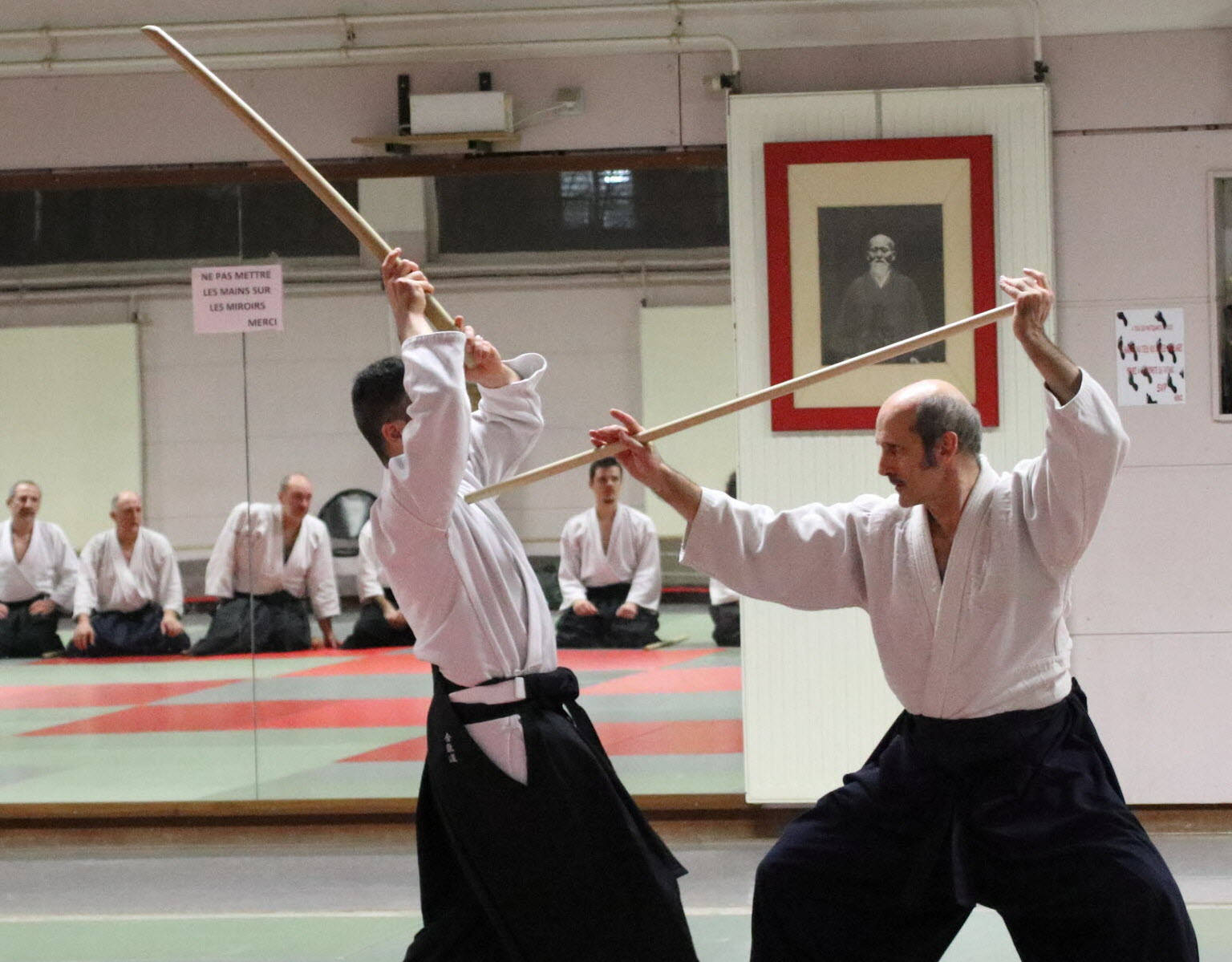 合氣道練習者於傳統道場持杖練習 壁紙 — 兩位身穿白色道服的武術家在道場中用木製木劍對打，學生們坐著觀看。