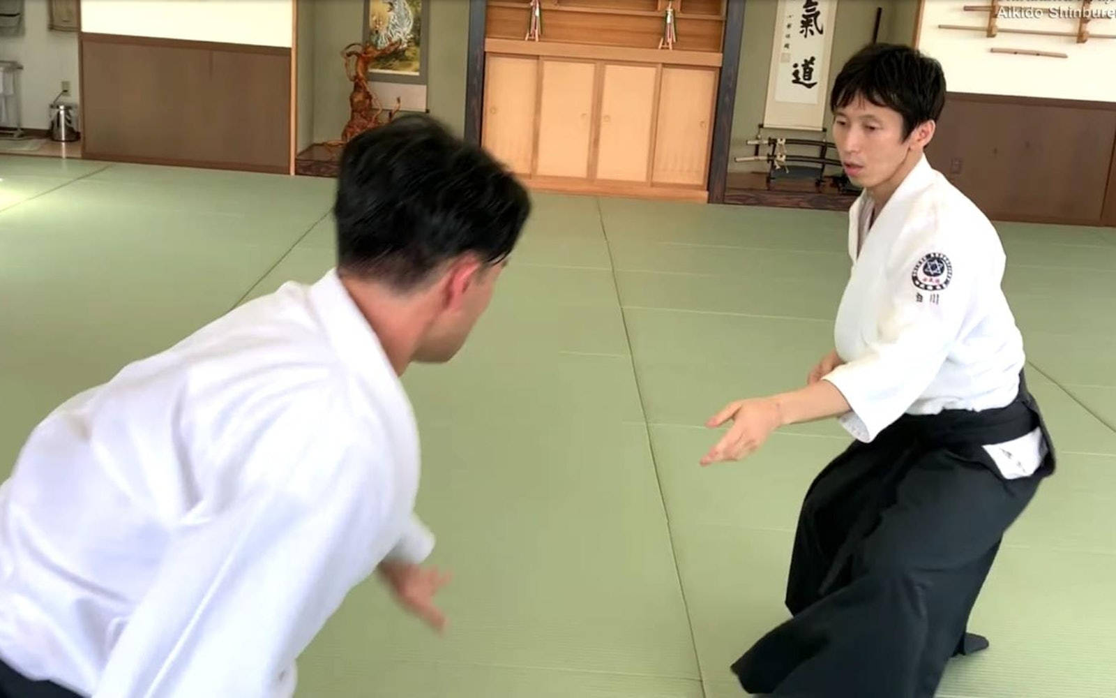 合氣道練習者於道場展示防禦姿勢 壁紙 — 兩名身穿白色道服嘅男子喺傳統道場嘅綠色榻榻米上練習合氣道，道場有木牆同書法。