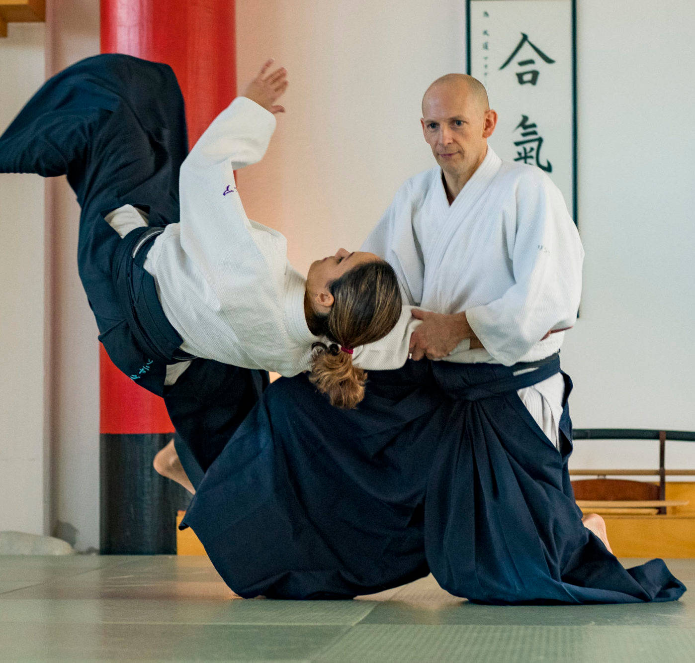 合氣道腰投技完美執行 壁紙 — 合氣道練習者在傳統道場的墊子上執行一個動態的投擲，牆上有書法。