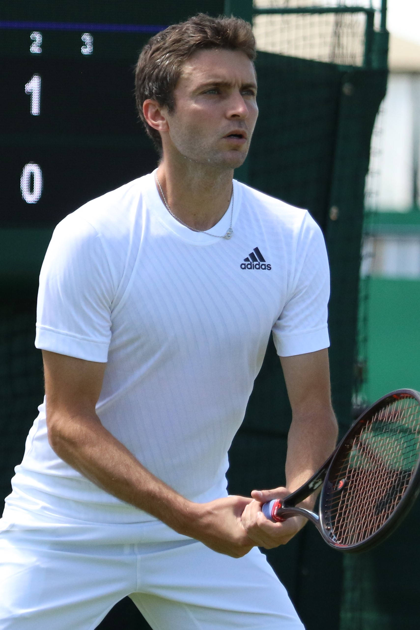吉勒斯·西蒙運動手機壁紙 壁紙 — 一位專注的男性網球選手穿著白色adidas裝備，在草地場上比賽中握著球拍，計分板清晰可見。