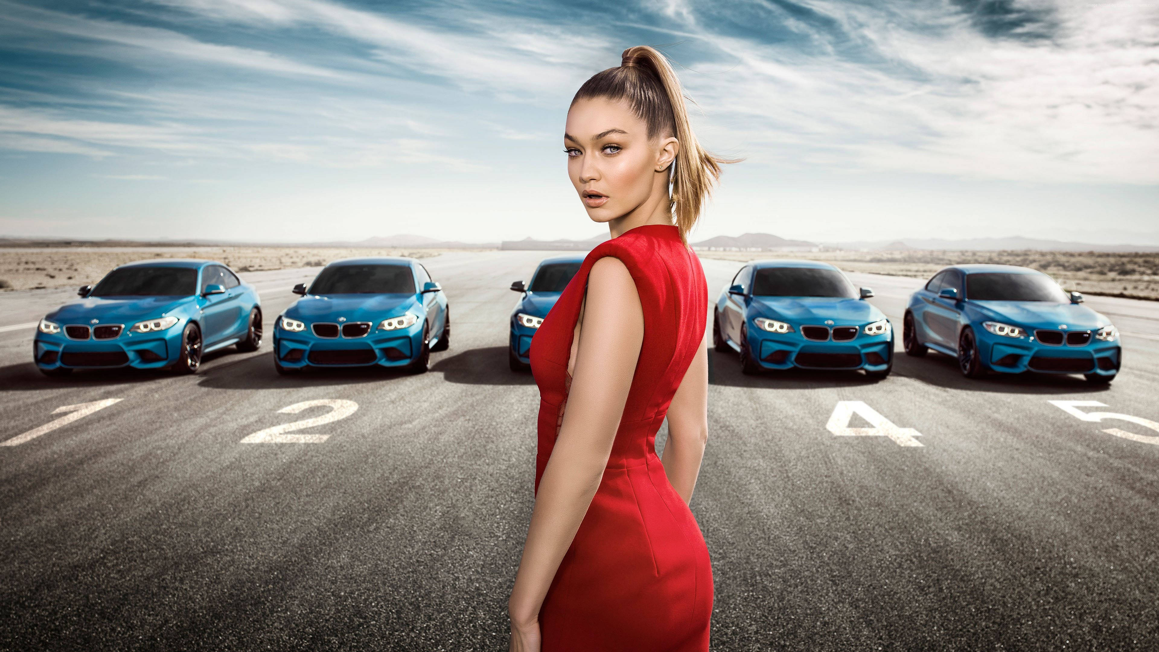 吉吉·哈迪德 與 BMW M2 車款 壁紙 — 一位身穿紅色連衣裙的女性自信地站在沙漠賽道上，五輛藍色寶馬跑車在她身後排成一排。