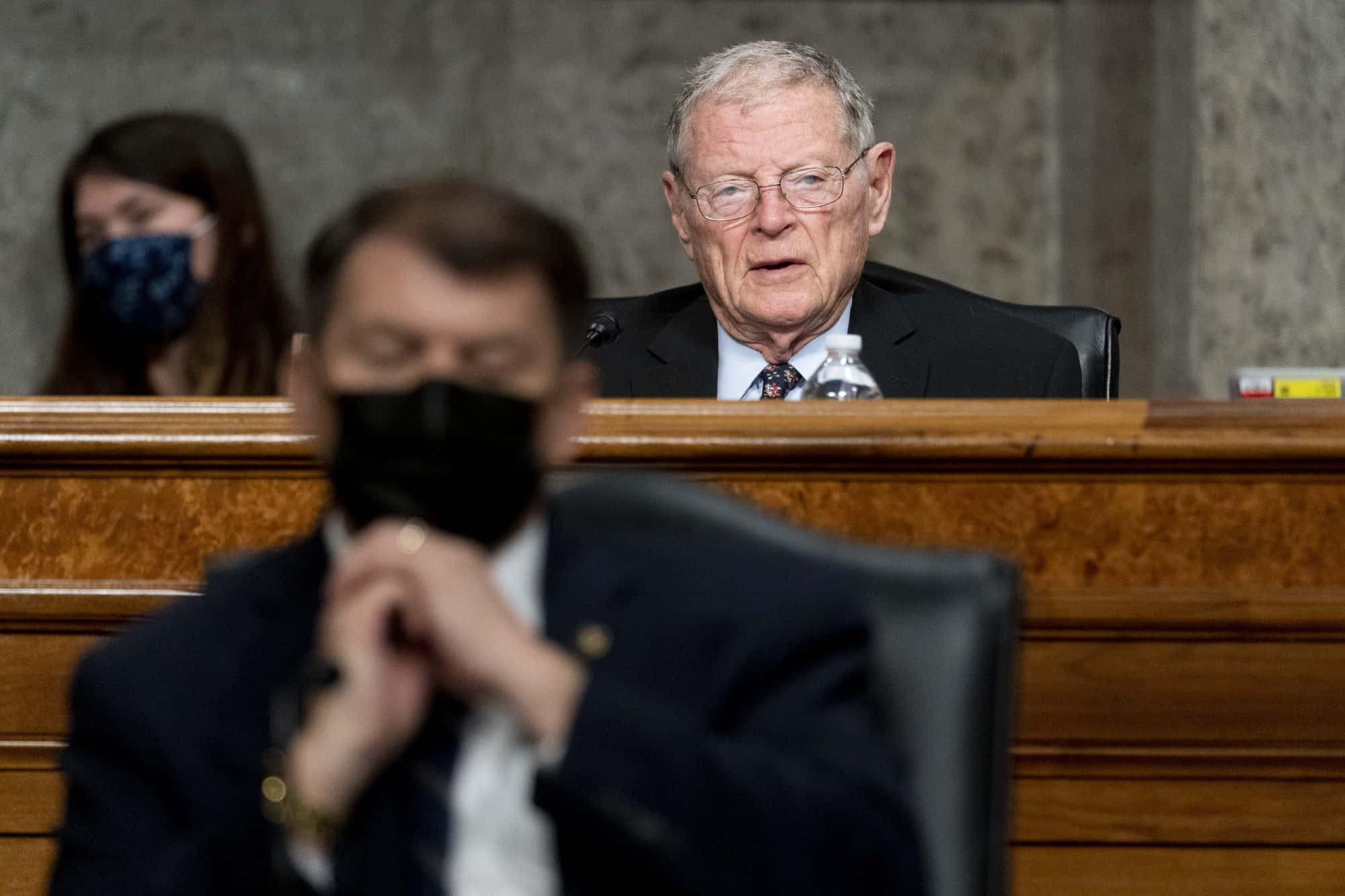 吉姆·因霍夫在會議中 壁紙 — 一位身穿西裝、戴眼鏡的年長男性在木製聽證桌前發言，背景中其他人戴著口罩。