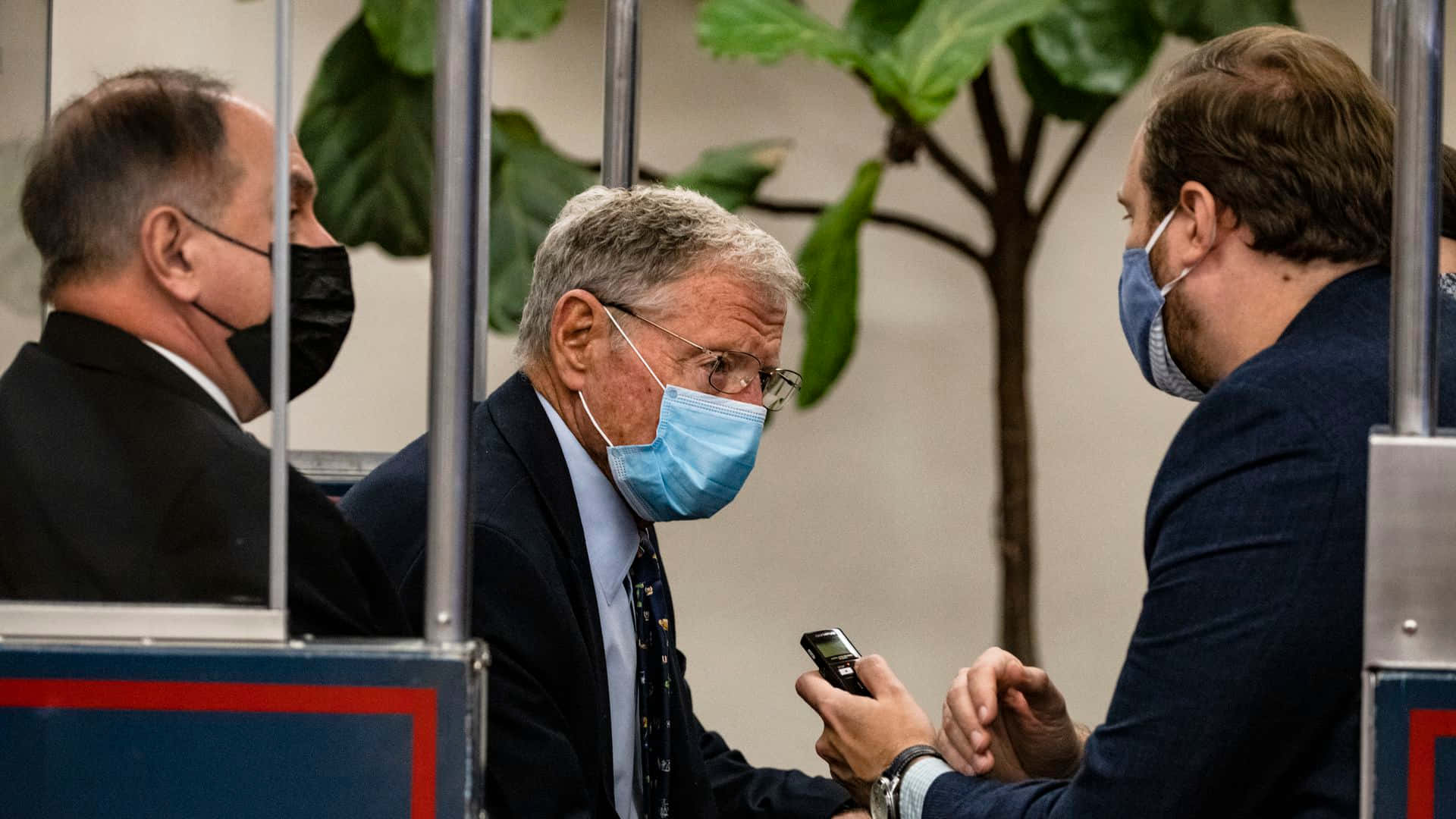 吉姆·因霍夫與記者對話 壁紙 — 三名身穿西裝、戴著口罩的男子在現代辦公室的大型琴葉榕後面，隔著玻璃隔板交談。