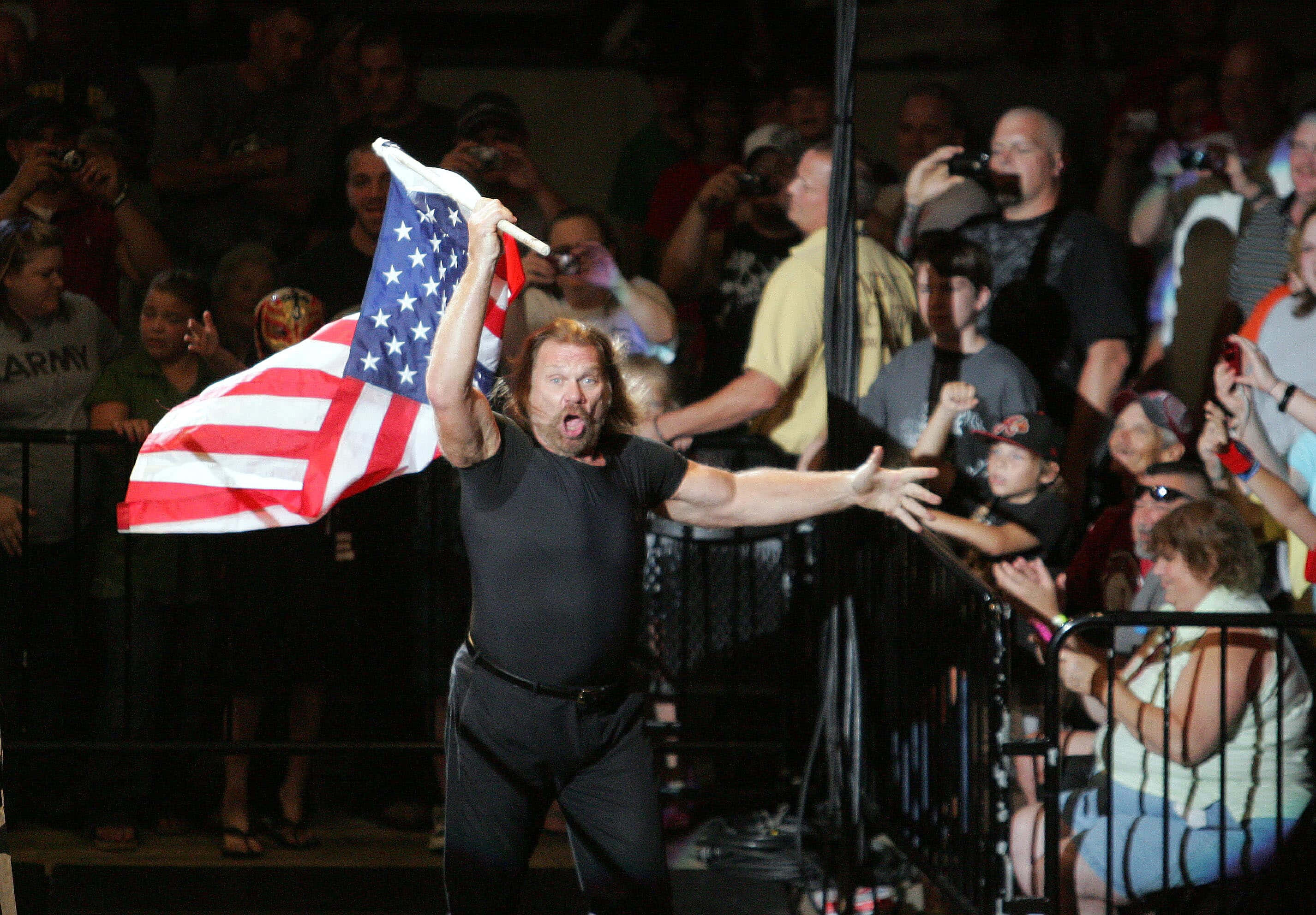 吉姆·達根參與WWE Raw世界巡迴賽 壁紙 — 一名肌肉發達的摔角手身穿黑色裝備，在擠滿觀眾的場館內揮舞美國國旗並咆哮。