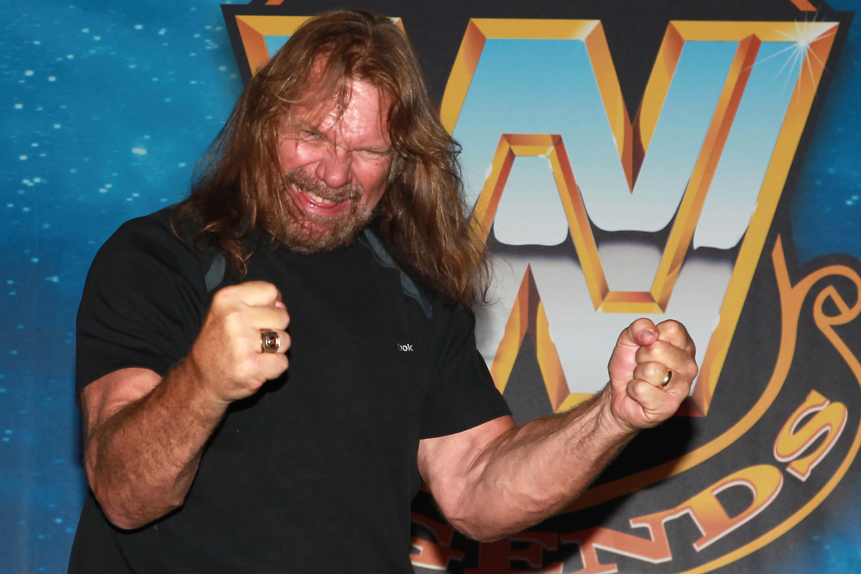 吉姆·達根雙手握拳慶祝 壁紙 — 長髮鬍鬚摔角手在WWE Legends標誌背景前揮舞拳頭。