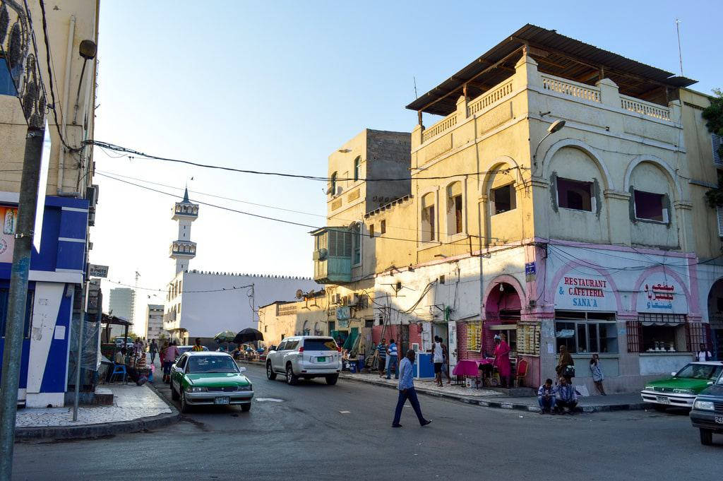陽光明媚的城市街道，有清真寺宣禮塔，人們走過商店和汽車。