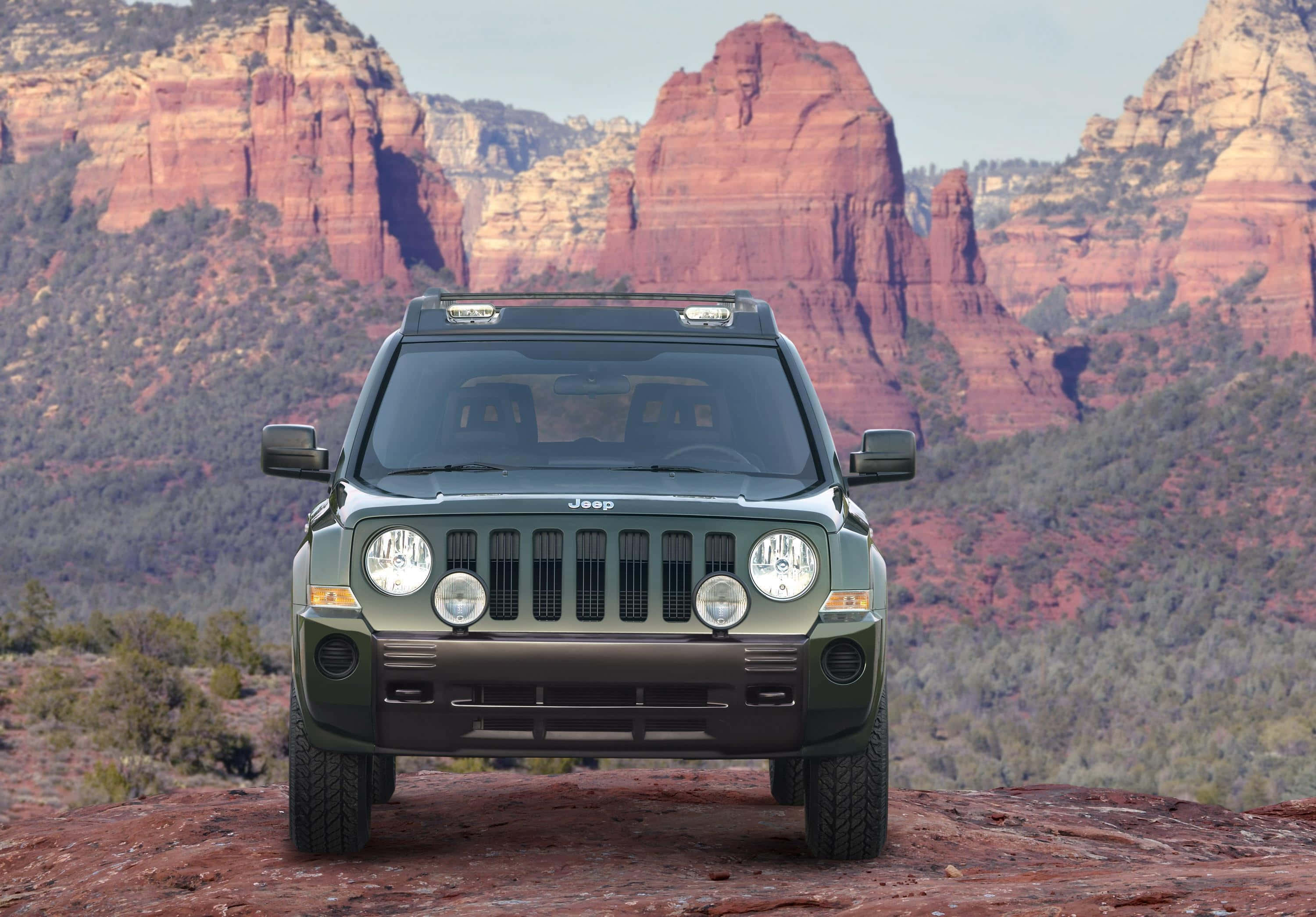吉普Patriot征服戶外風景 壁紙 — 一輛綠色Jeep Wrangler在晴朗天空下，雄偉地矗立在紅色岩石地形上，背後是高聳的深紅色峽谷牆壁。