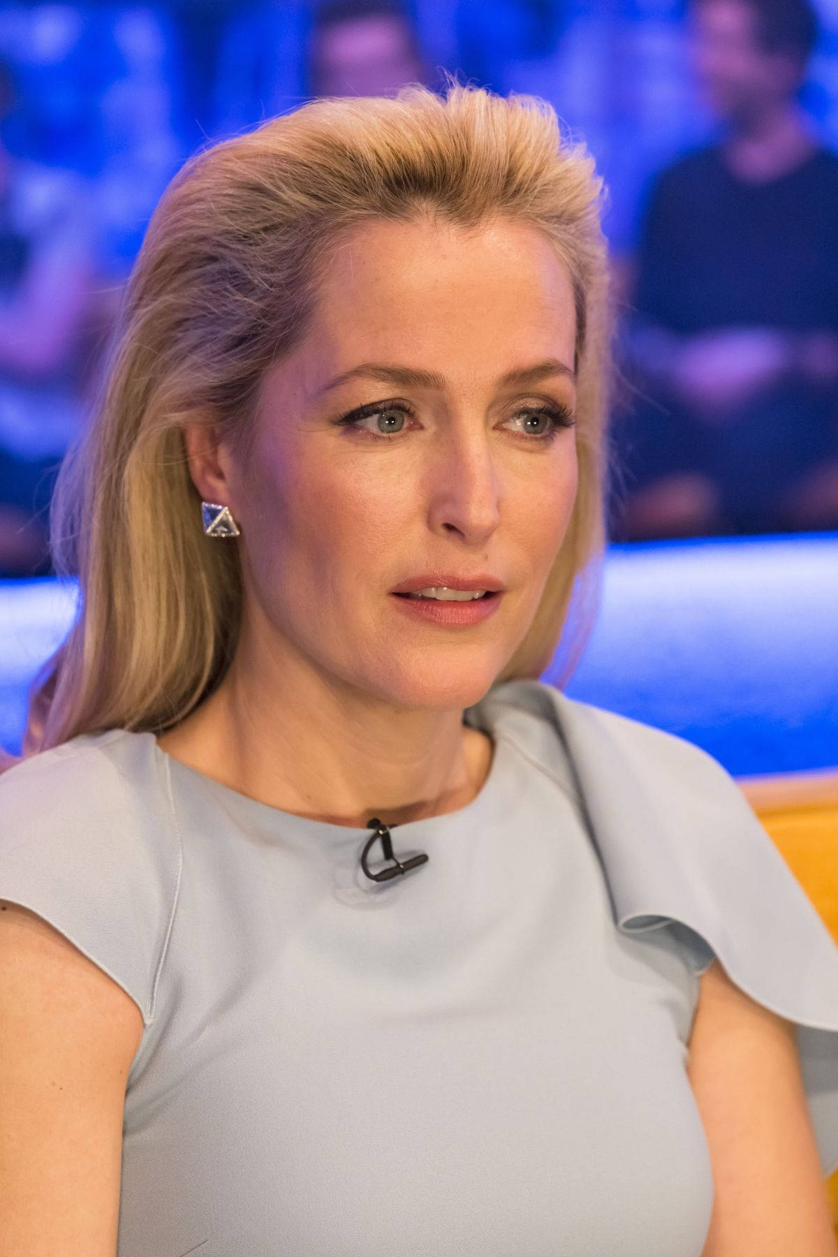 吉蓮·安德森 Jonathan Ross 節目 壁紙 — 一位金髮女子穿著淺灰色連衣裙，坐在藍色燈光、背景有模糊觀眾的攝影棚中。