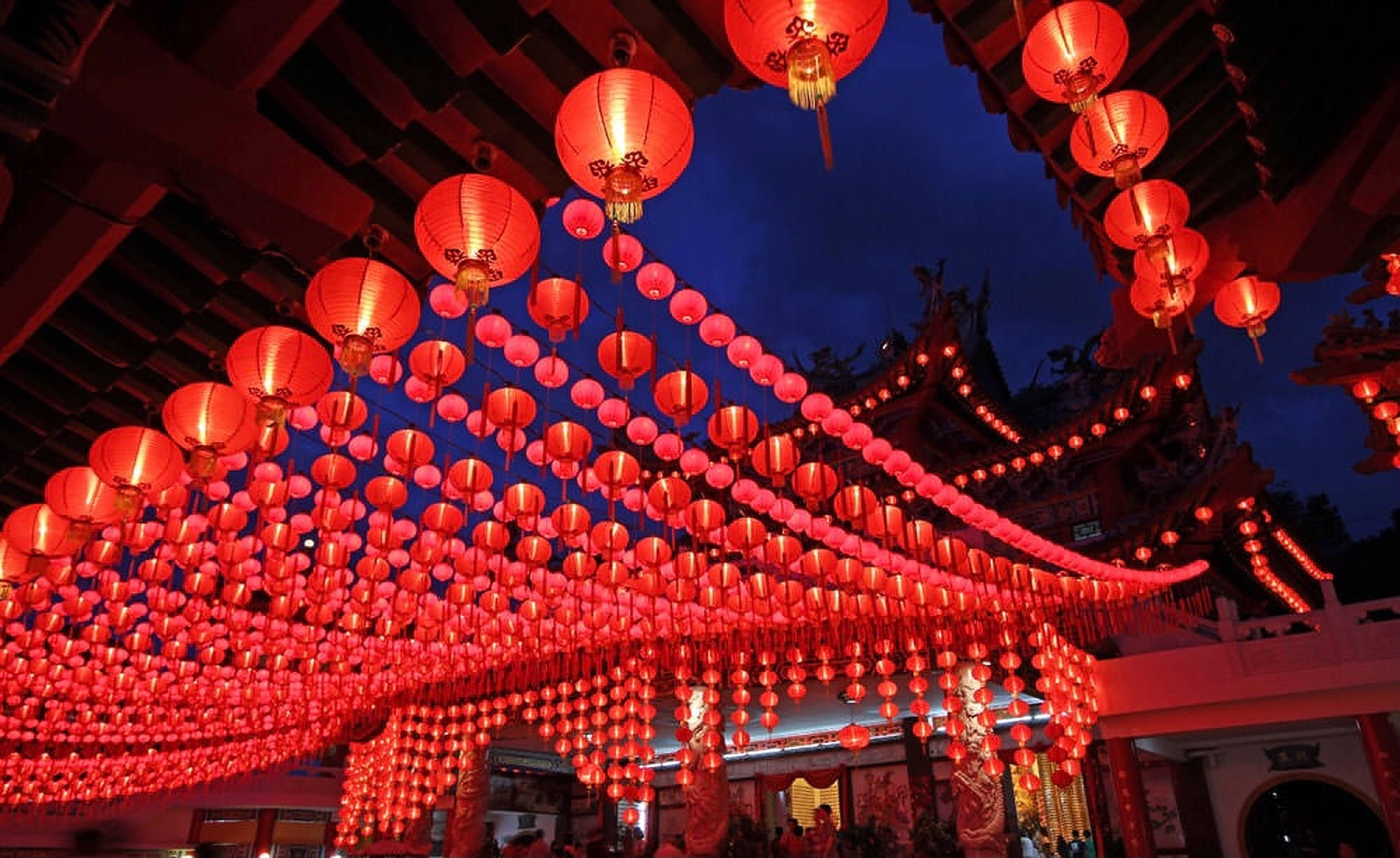 吉隆坡夜間燈籠盛景 壁紙 — 數千盞發光的紅色燈籠如瀑布般懸掛在傳統寺廟屋頂上，映襯著深藍色的暮色天空。