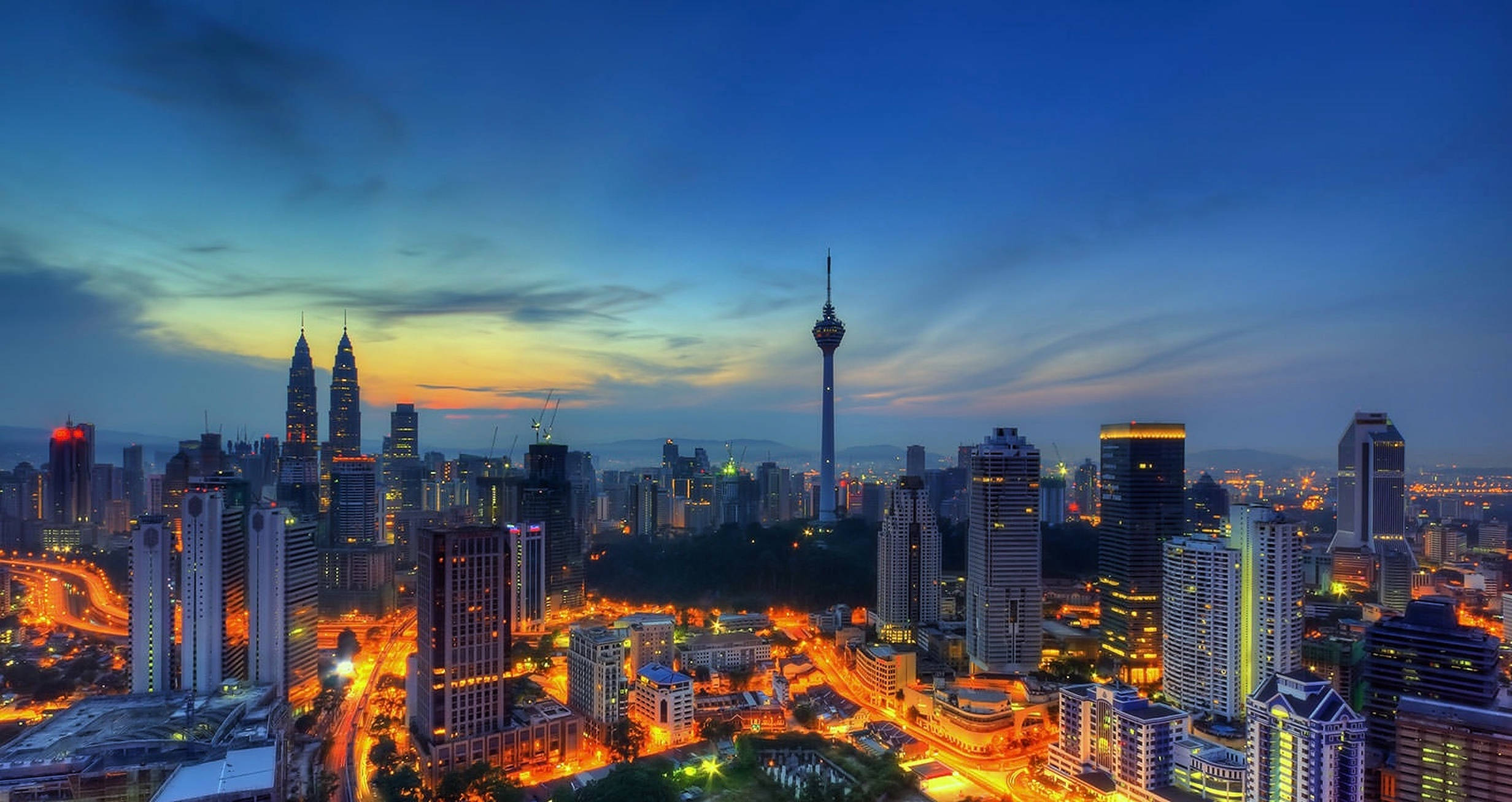 吉隆坡市中心夜景 壁紙 — 黃昏時分充滿活力的城市景觀，閃爍的摩天大樓和蜿蜒的道路在漸變藍色天空下。