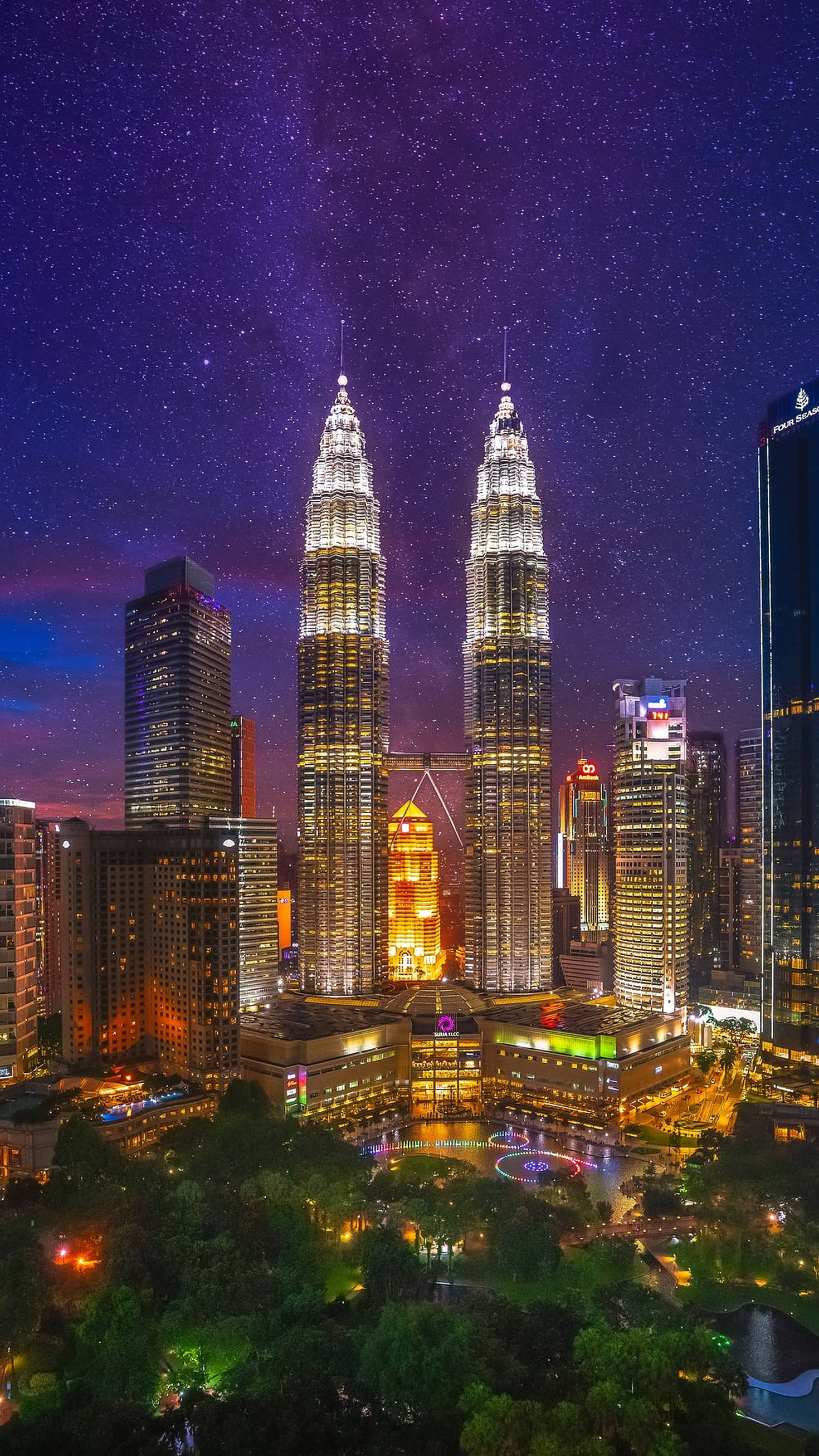 吉隆坡繁星夜景 壁紙 — Twin skyscrapers glow under a starry night sky above a vibrant cityscape with glowing lights and lush greenery below.