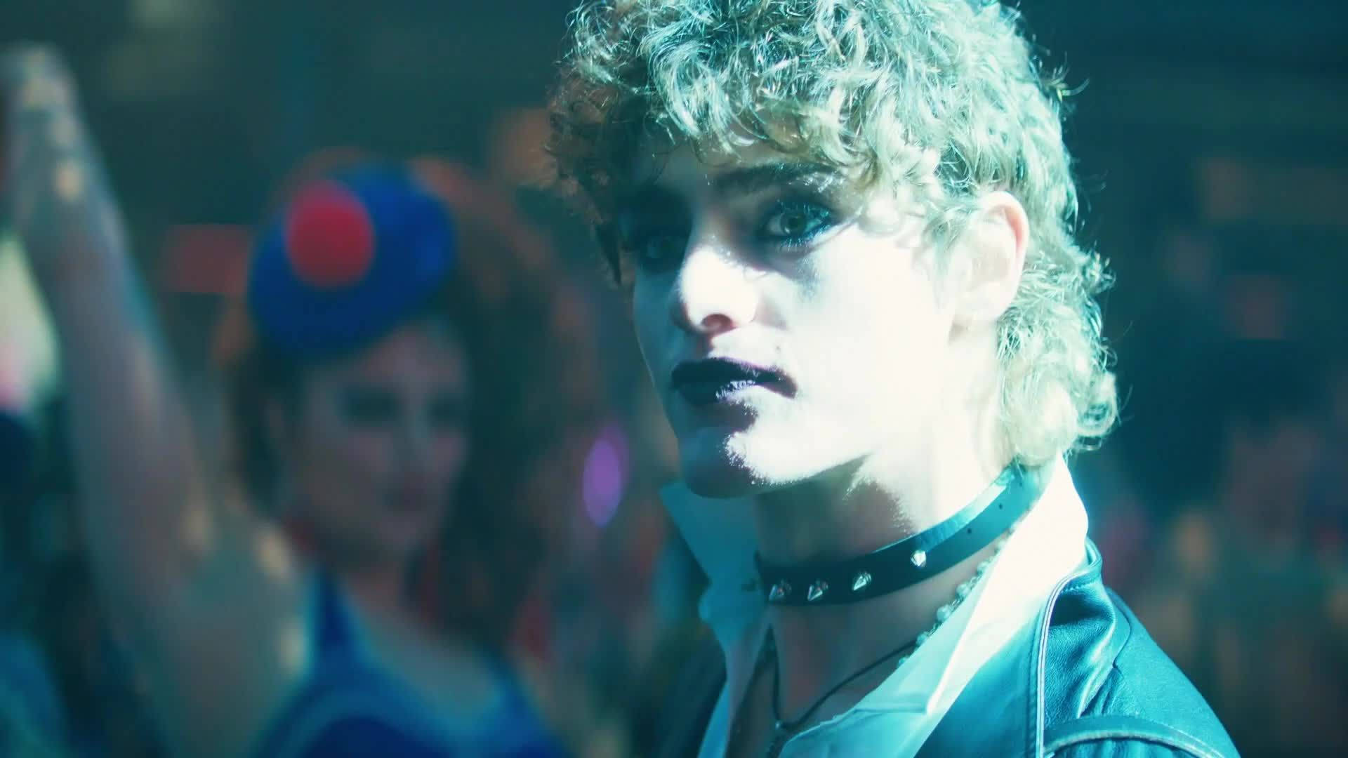 《同志亦凡人》朱利安夜店風采 壁紙 — 一個擁有捲曲金髮和深色唇膏的人，在燈光昏暗、彩色燈光和模糊舞者的俱樂部中強烈凝視。