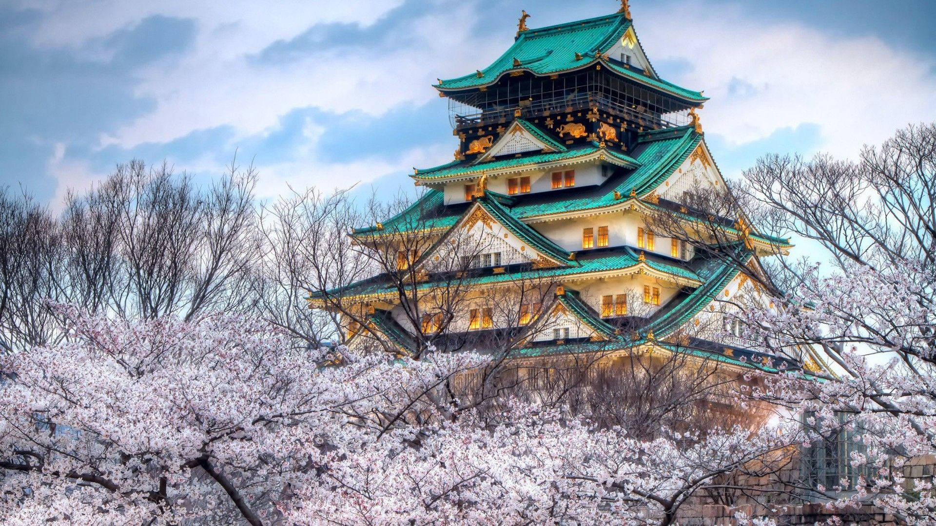 名古屋城冬季風景 壁紙 — 一座雄偉的日本城堡，擁有綠色屋頂和發光的窗戶，在陰雲密布的天空下，從盛開的粉紅色櫻花後方升起。