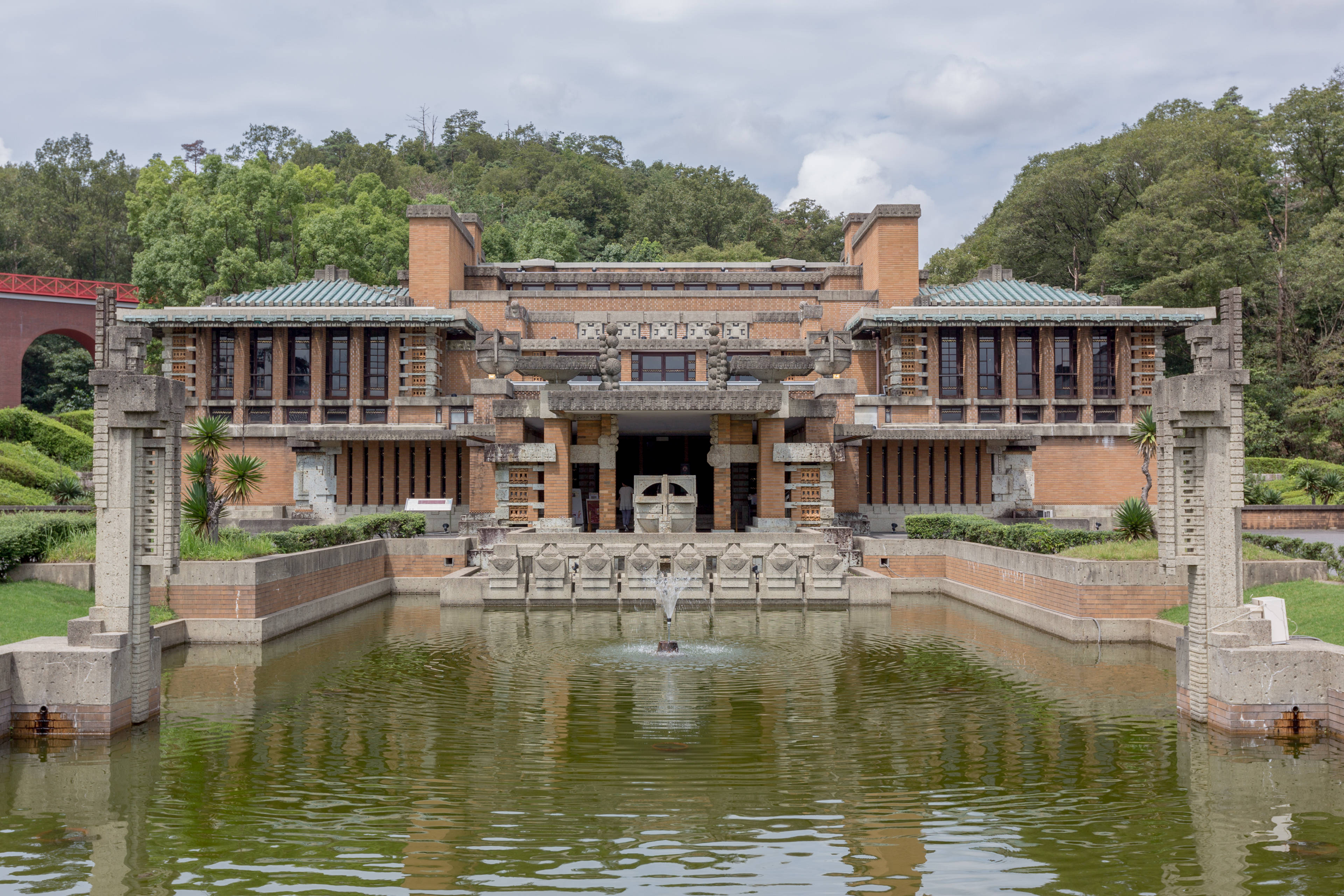 名古屋明治村博物館 壁紙 — 一座宏偉的草原風格豪宅，採用赤陶磚和綠色瓦片屋頂，倒映在寧靜的泳池中，中央噴泉被茂密的綠植環繞。