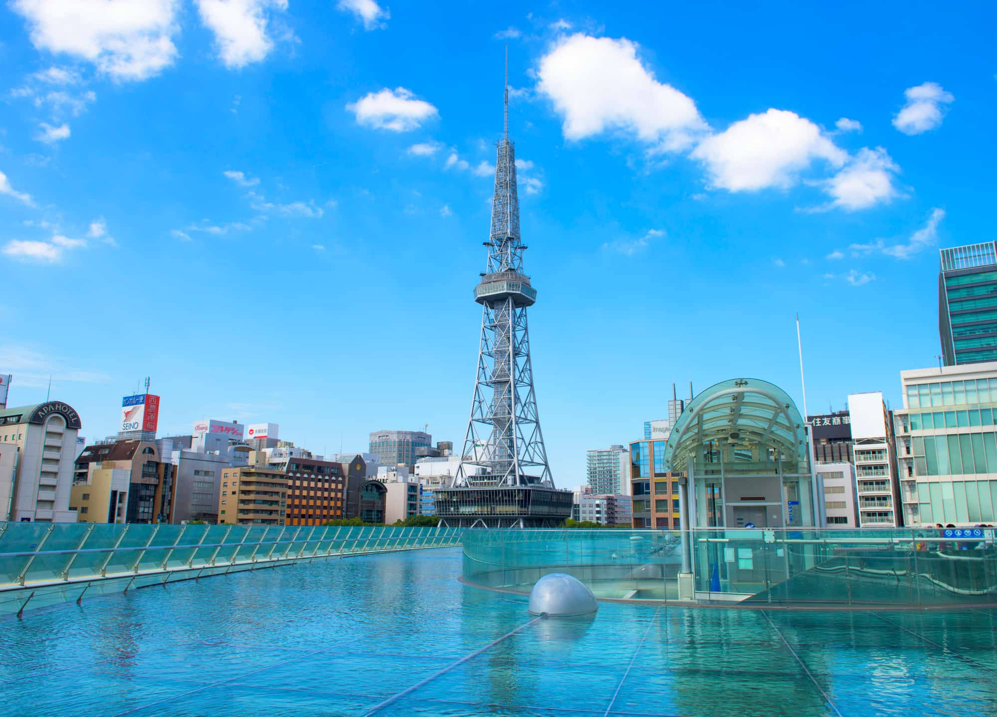 名古屋未來電視塔 壁紙 — 明亮的藍天白雲俯瞰著現代城市景觀，包括一座高鋼塔和一個玻璃屋頂建築，旁邊有一個反射池。