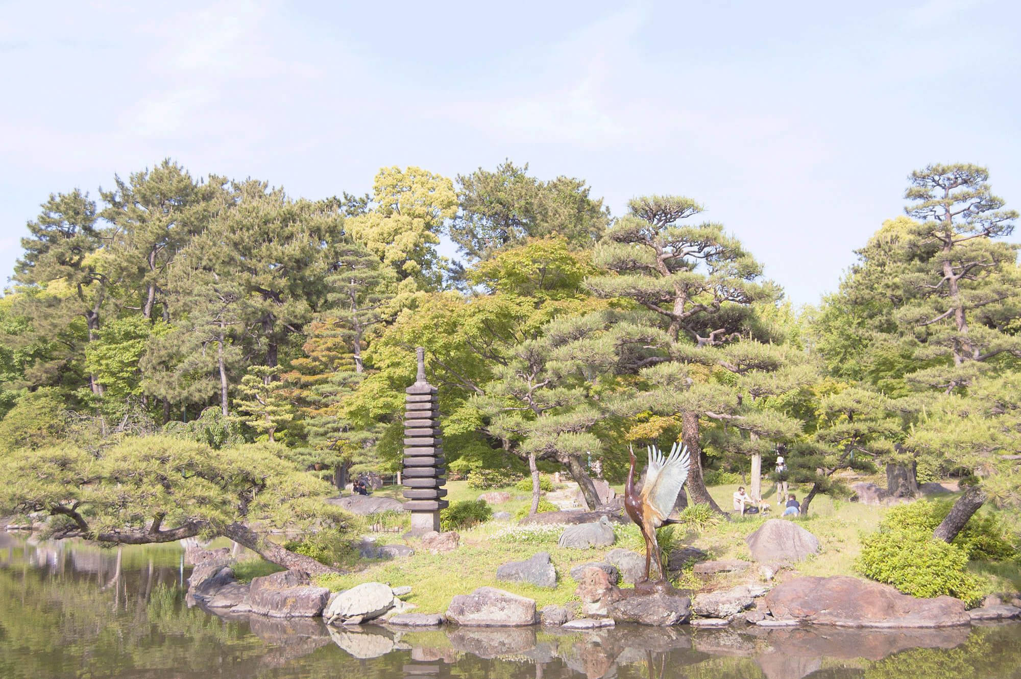 名古屋禪園 壁紙 — 一個寧靜的日本花園池塘倒映著石塔和鶴雕像，周圍環繞著茂密的綠樹，天空明亮。