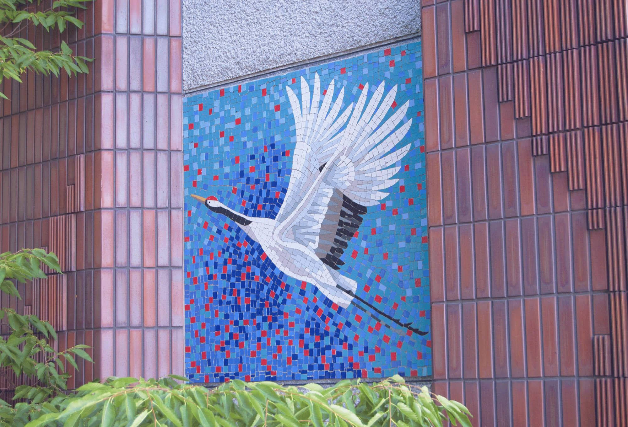 名古屋鶴舞公園鶴形藝術牆 壁紙 — 一隻充滿活力的馬賽克鶴在飛行中，點綴著被綠色植物環繞的現代建築外牆。