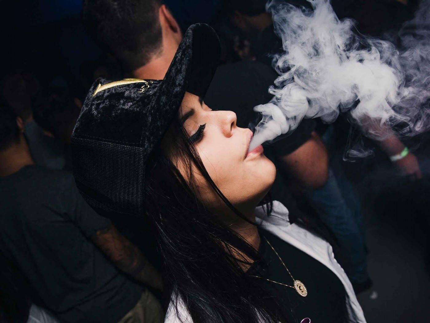吐煙的態度女孩 壁紙 — 一位戴黑色帽子的年輕女性在昏暗擁擠的場所呼出濃厚的白色蒸氣。