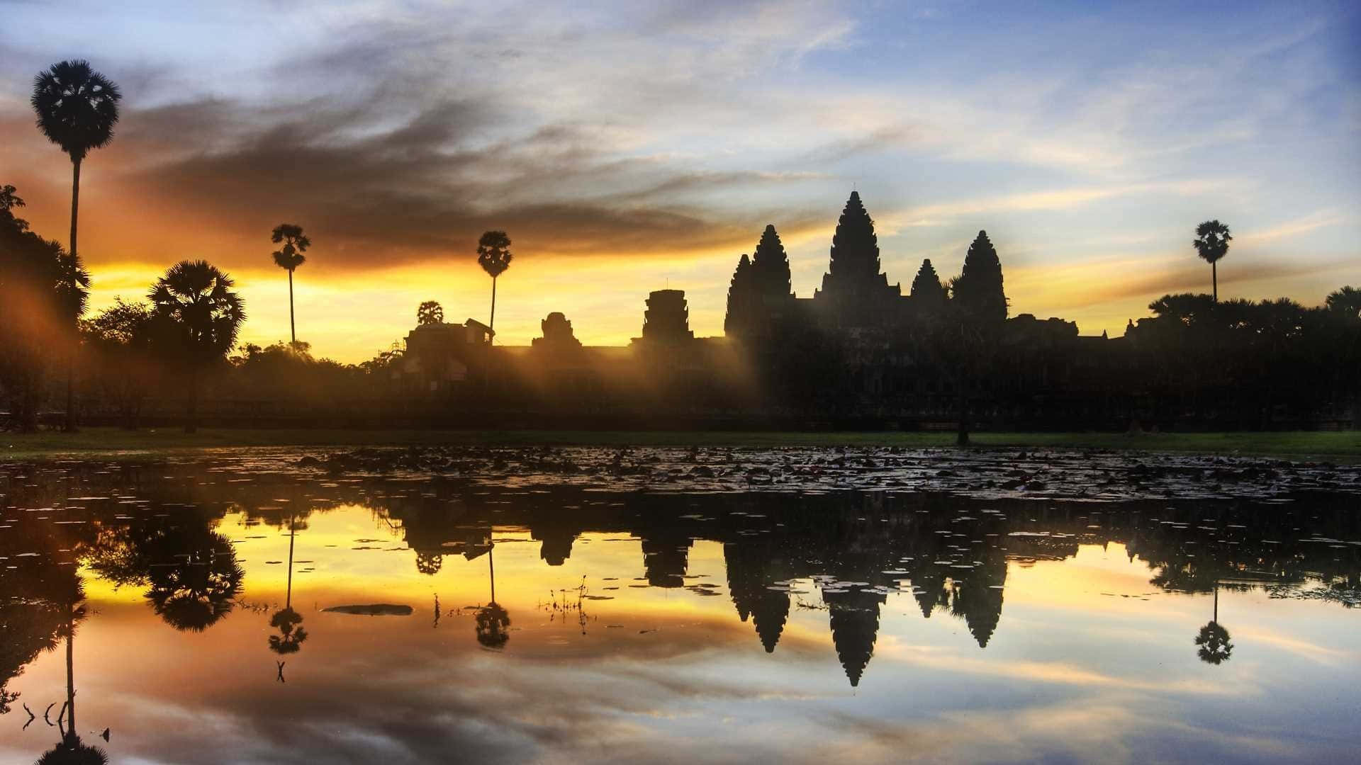 Thailand 桌布 壁紙 — 吳哥窟寺廟的剪影倒映在平靜的水面上，金色的夕陽光芒灑滿天空和棕櫚樹。