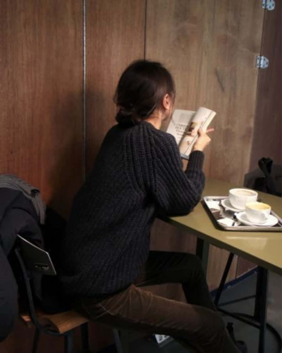 咖啡店內閱讀的女學生 壁紙 — 一個穿著深色針織毛衣的人在一間舒適的咖啡館裡，在木桌上放著兩杯咖啡，正在閱讀一本書。