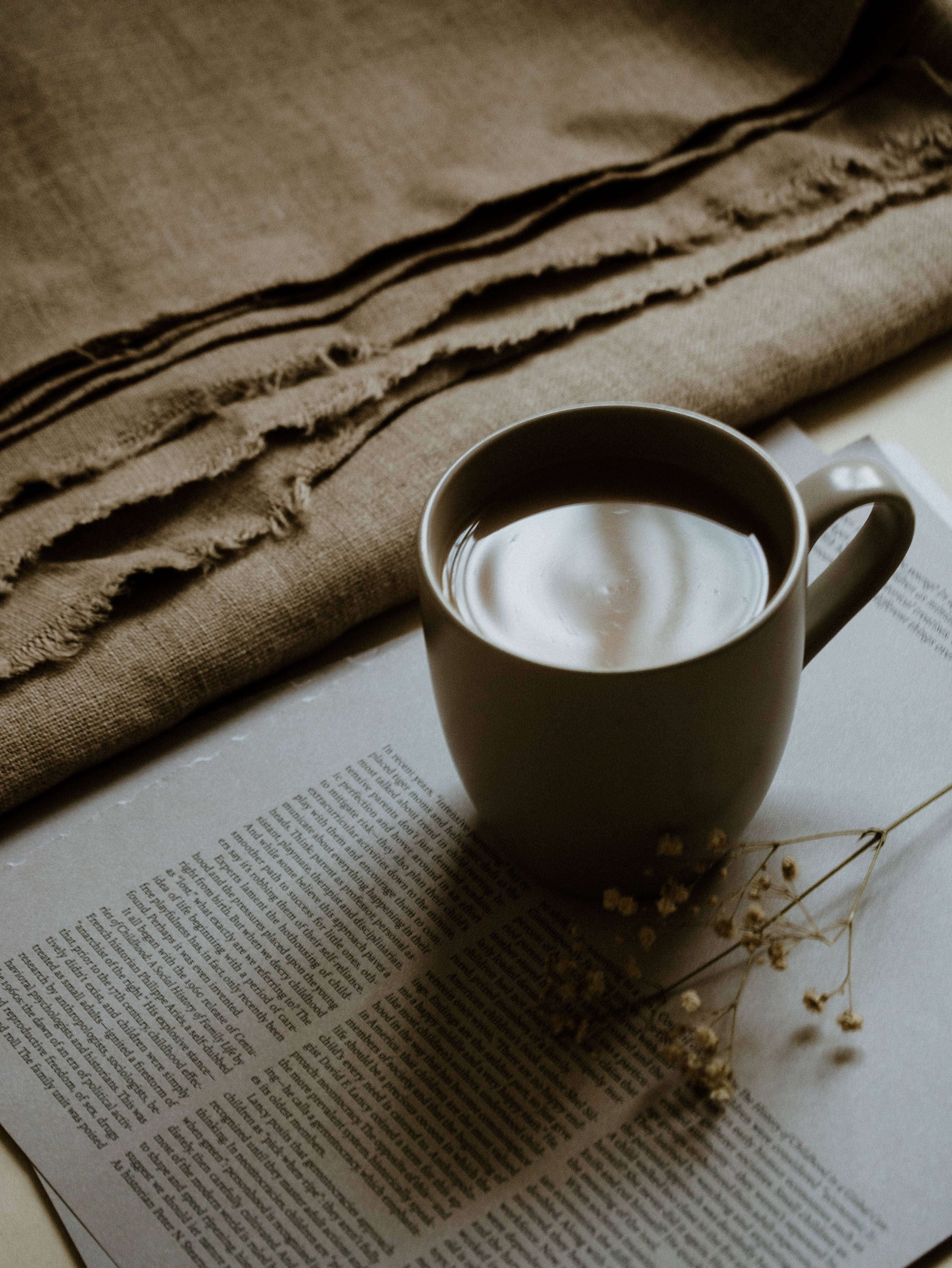 咖啡杯下的雜誌 壁紙 — 一杯冒煙的咖啡放在一本打開的書上，旁邊有乾花和一塊有紋理的米色布料，在柔和自然光下。