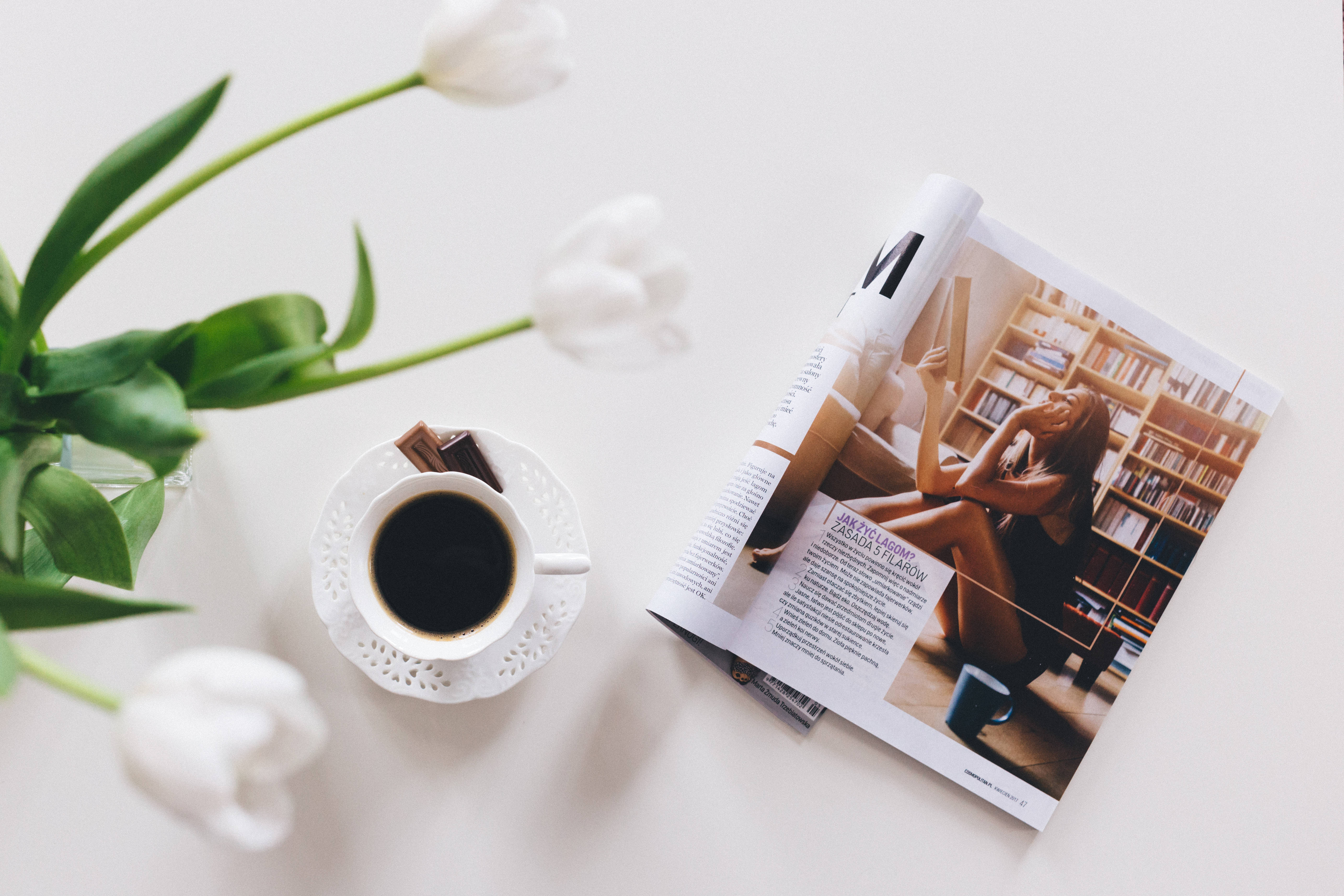 咖啡與花朵旁的雜誌 壁紙 — 一個寧靜的平鋪構圖，包含白色鬱金香、一杯黑咖啡和一本打開的雜誌，放在潔白的表面上。