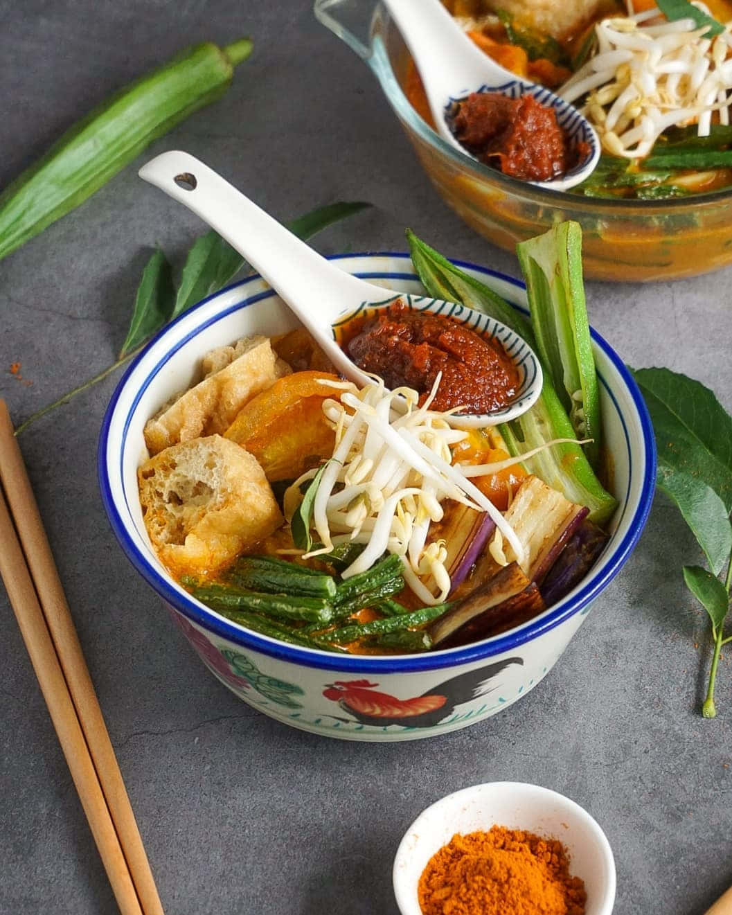 咖喱叻沙食材平鋪擺設 壁紙 — 一碗冒著蒸氣的東南亞風味麵湯，上面放有豆芽、秋葵、豆腐和辣椒醬。