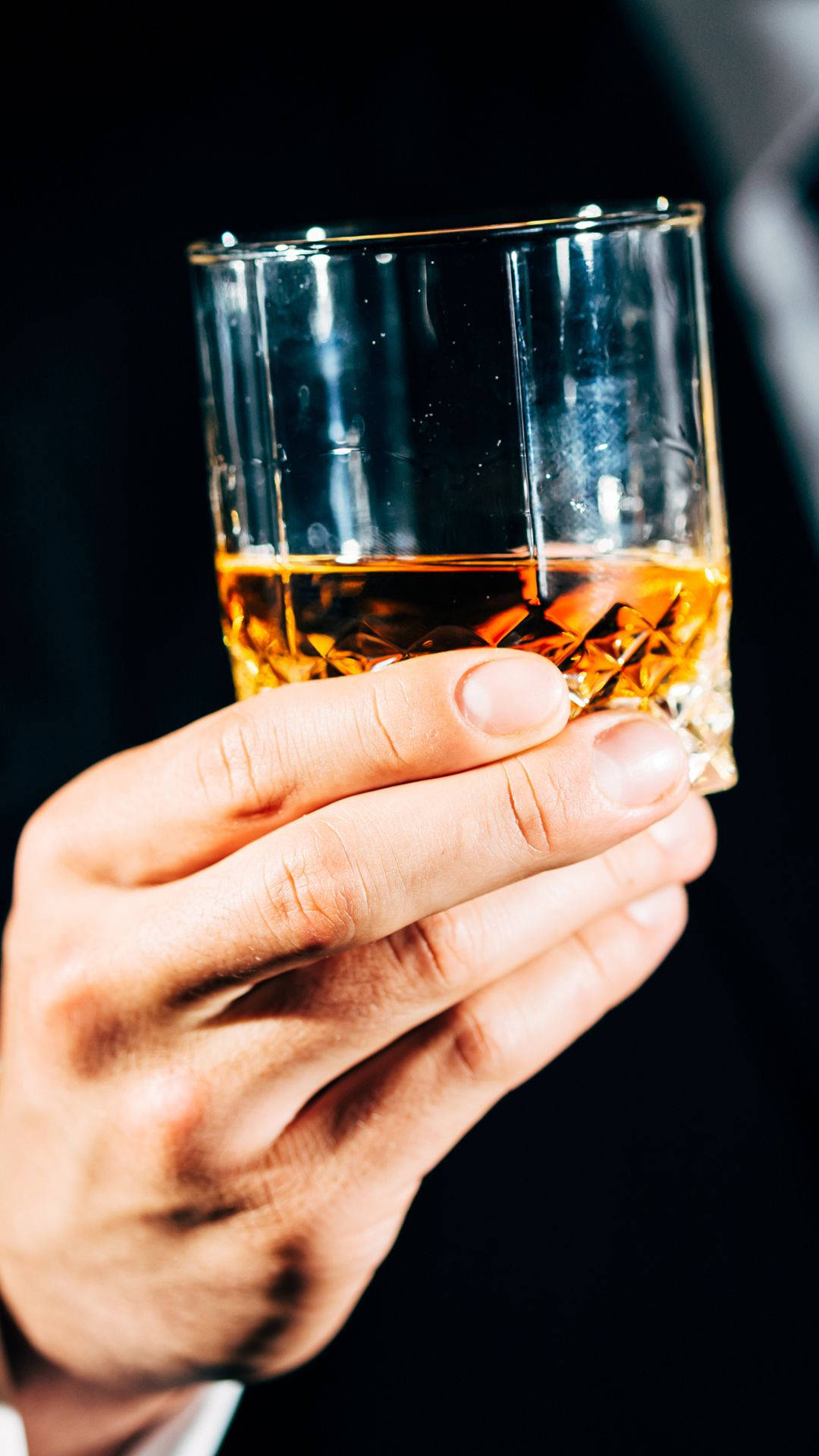 品味人生時刻，舉杯有格調 壁紙 — 一隻手拿著一個水晶杯，裡面裝著琥珀色的威士忌，背景昏暗，光線柔和。
