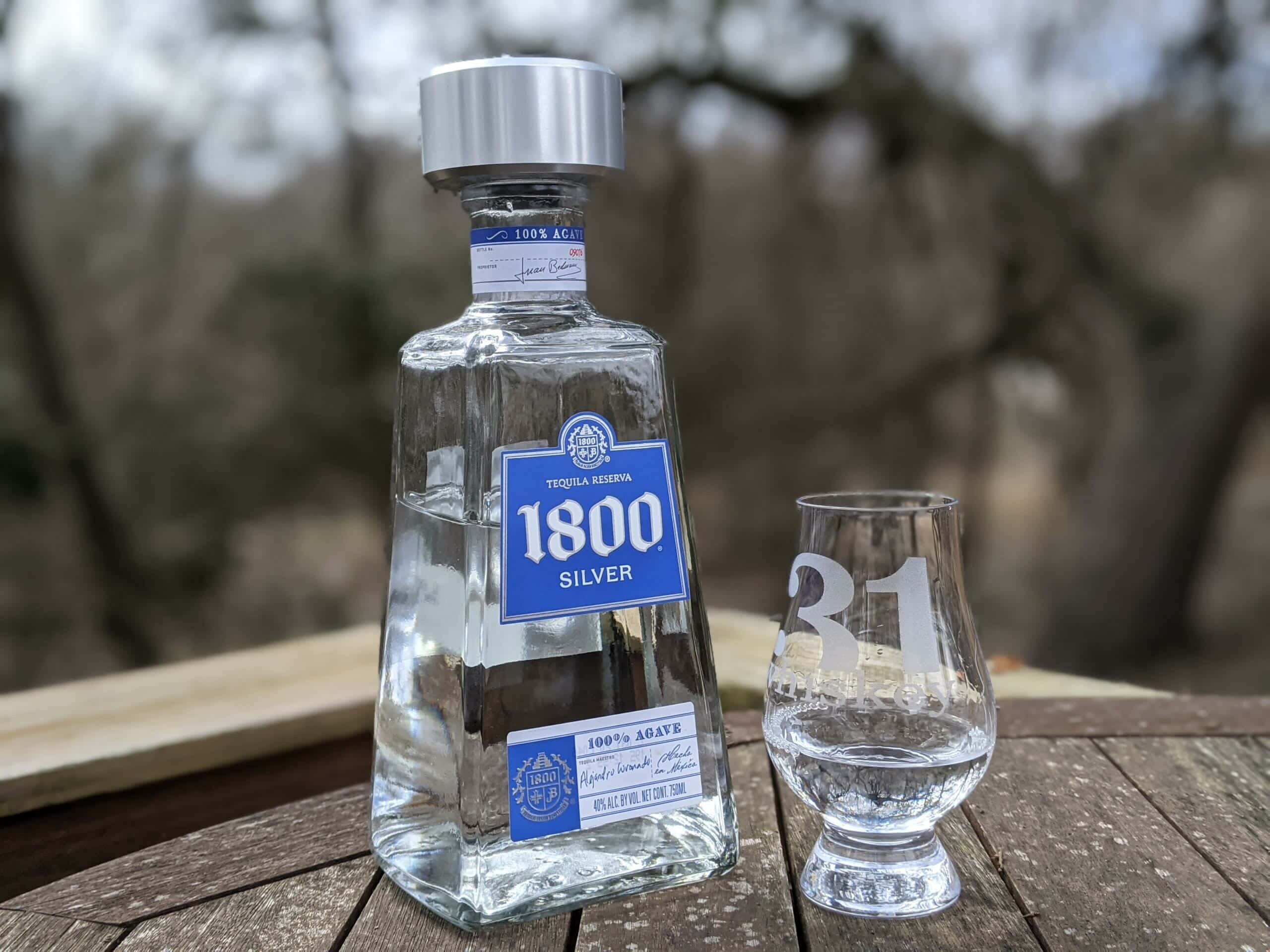 品味最精緻——1800 蒂奎拉銀標酒瓶 壁紙 — 一個透明的1800 Silver龍舌蘭酒瓶和一個刻有「31」的玻璃杯放在戶外一張風化的木桌上。