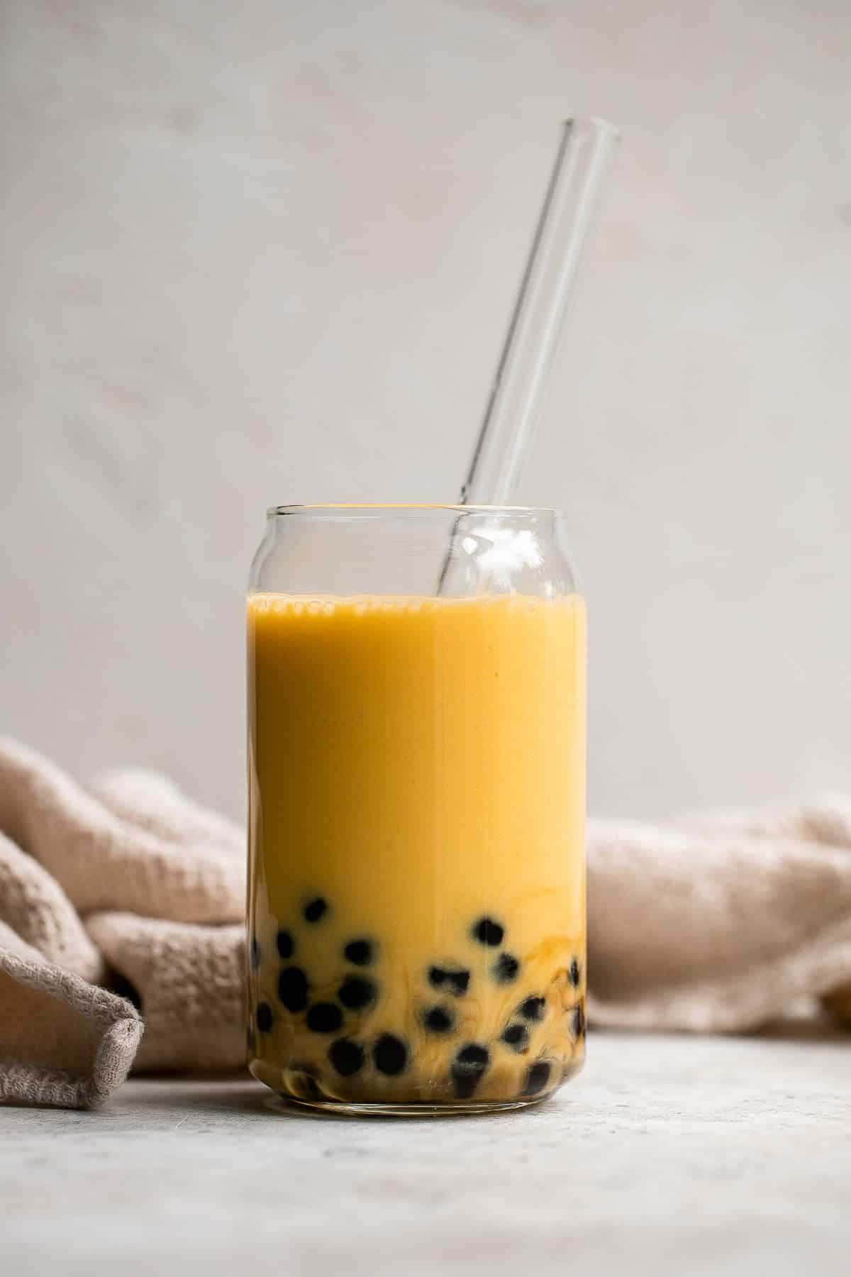 品味珍珠奶茶的清爽滋味 壁紙 — 一個玻璃罐中的鮮黃色珍珠奶茶，配有黑色木薯珍珠和透明吸管，背景是柔軟的米色布料。