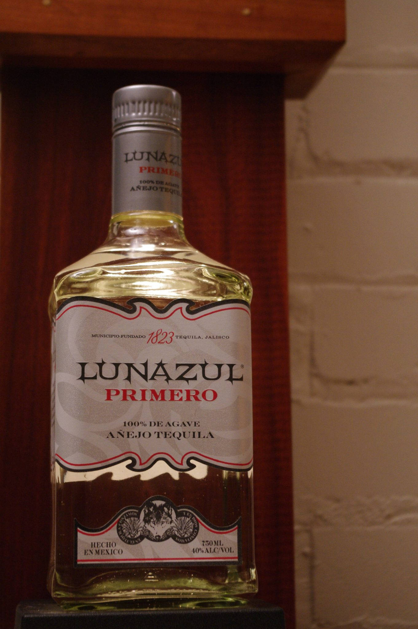 品味真實風味：Lunazul Primero 蒂克酒 壁紙 — 一瓶Lunazul Primero Añejo龍舌蘭酒放在一個磚牆和木框的架子上。