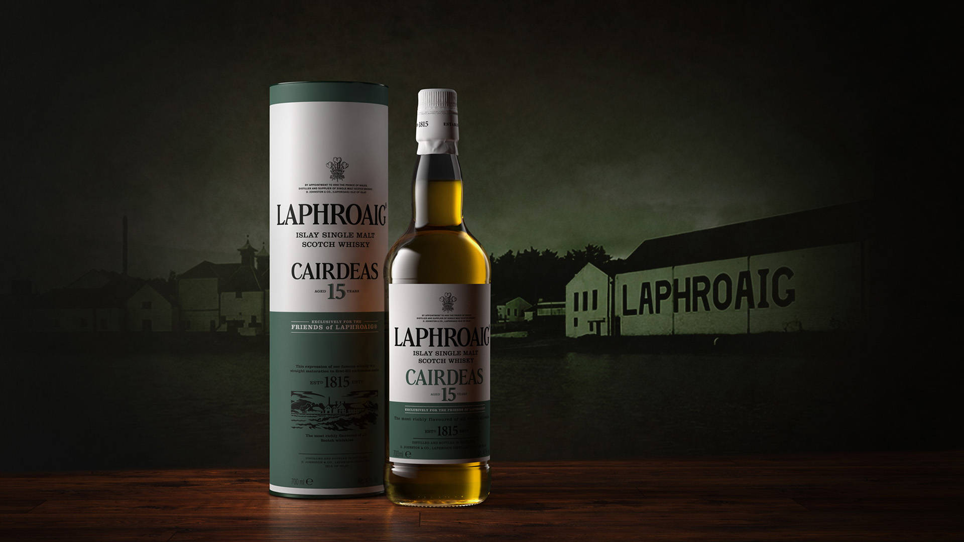 Laphroaig Cairdeas 15 年威士忌酒瓶和管狀包裝放在木桌上，背景是蒸餾廠。