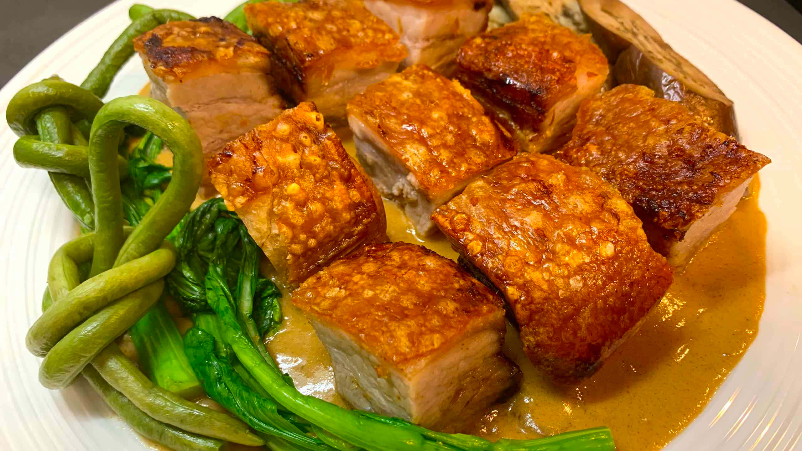 品嚐正宗菲律賓菜式——酥脆炸豬腩配濃郁 Kare-Kare 醬 壁紙 — 金黃酥脆的豬腩肉塊在濃郁的棕色醬汁中閃閃發光，旁邊是鮮綠色的白菜和四季豆，盛在白色碟子上。