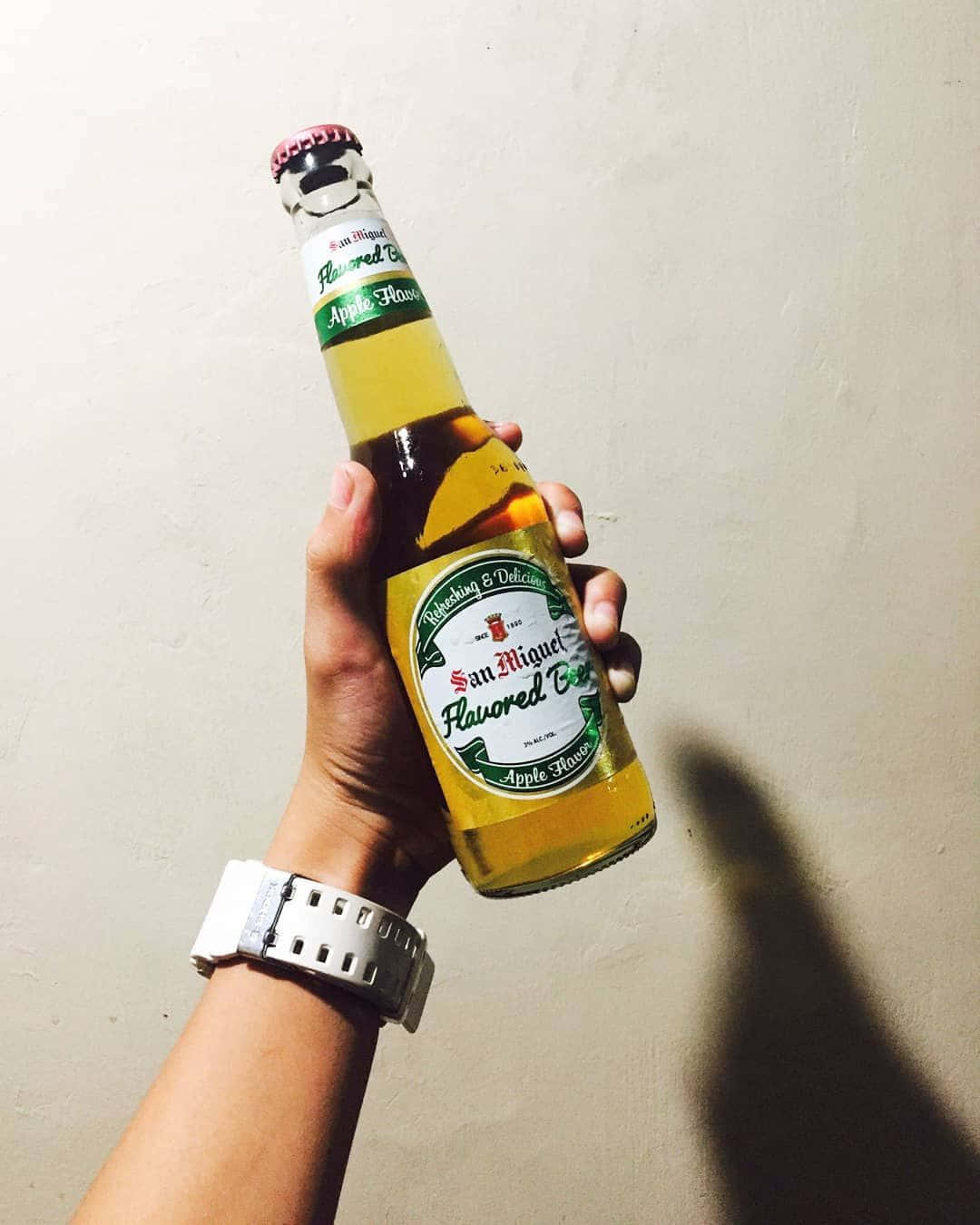 品嚐清爽啤酒 壁紙 — 一隻手拿著San Miguel蘋果味啤酒瓶靠著素色牆壁，手腕上戴著手錶。