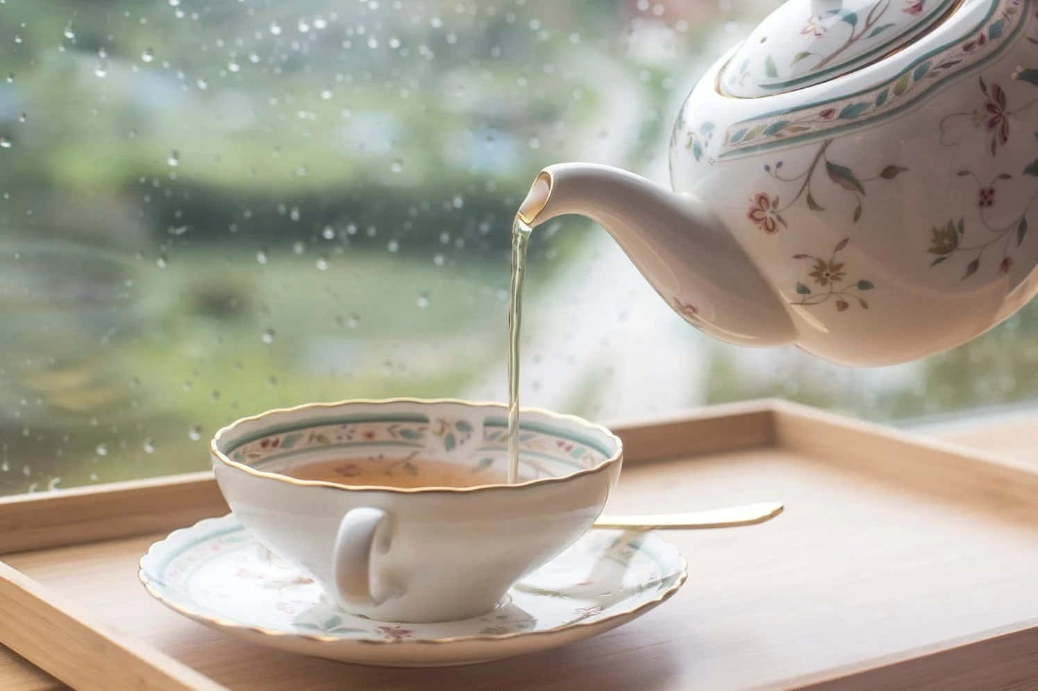 從花卉茶壺中倒出的冒煙茶，倒入一個相配的杯子中，杯子放在一個木托盤上，旁邊是下雨的窗戶。
