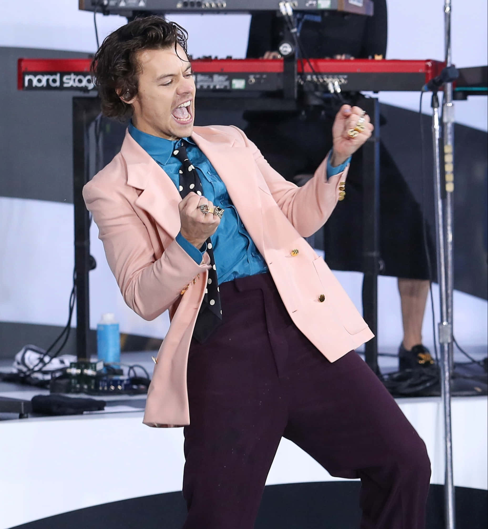 哈利·史蒂爾斯 2021 壁紙 — Harry Styles 身穿粉紅色西裝外套和紫色褲子，佩戴多枚戒指和一條波點領帶，在舞台上充滿活力地表演。